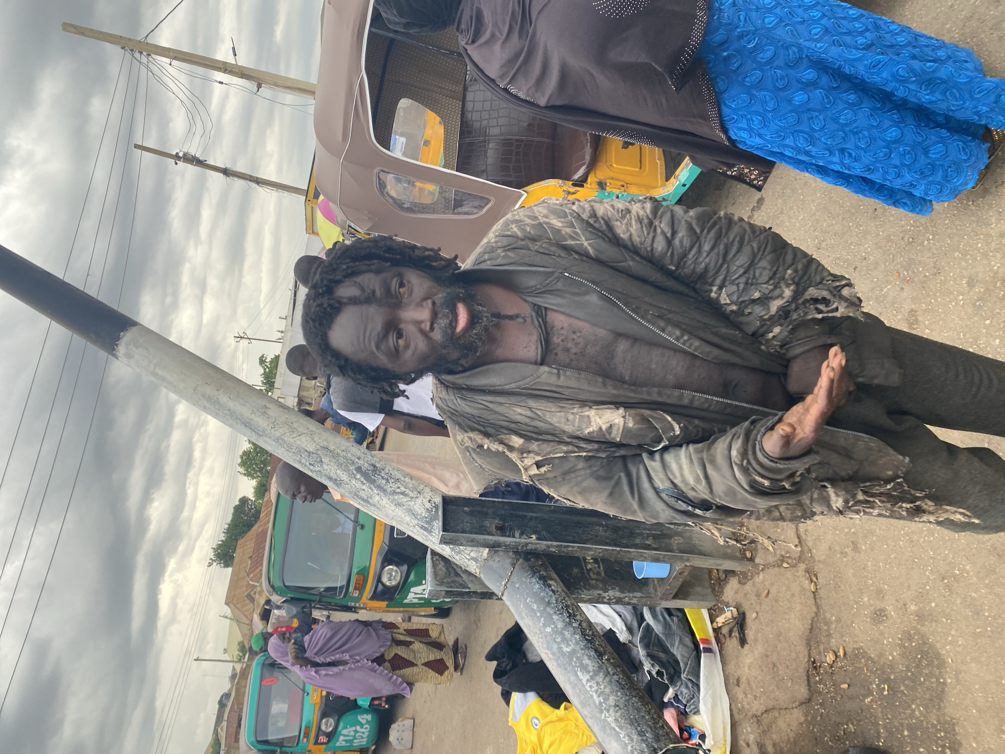 The scene is an outdoor setting in Gura Top, Nigeria. A man with dreadlocks and wearing tattered clothing is centrally positioned, leaning towards the viewer. His skin appears darkened, and he has a disheveled appearance.
Behind the man is a brown and yellow auto rickshaw (tricycle). To the right, a person in a black top and a bright blue patterned skirt is partially visible. In the background, other people are present, along with additional auto rickshaws, some green and some purple. One of the vehicles has a license plate that appears to read "PTA 1126-6".
A black and silver pole is in the foreground, partially obscuring the view. Litter and objects are scattered on the ground near the man. Overhead, power lines are visible against a cloudy sky. The environment appears urban, likely a street or marketplace.