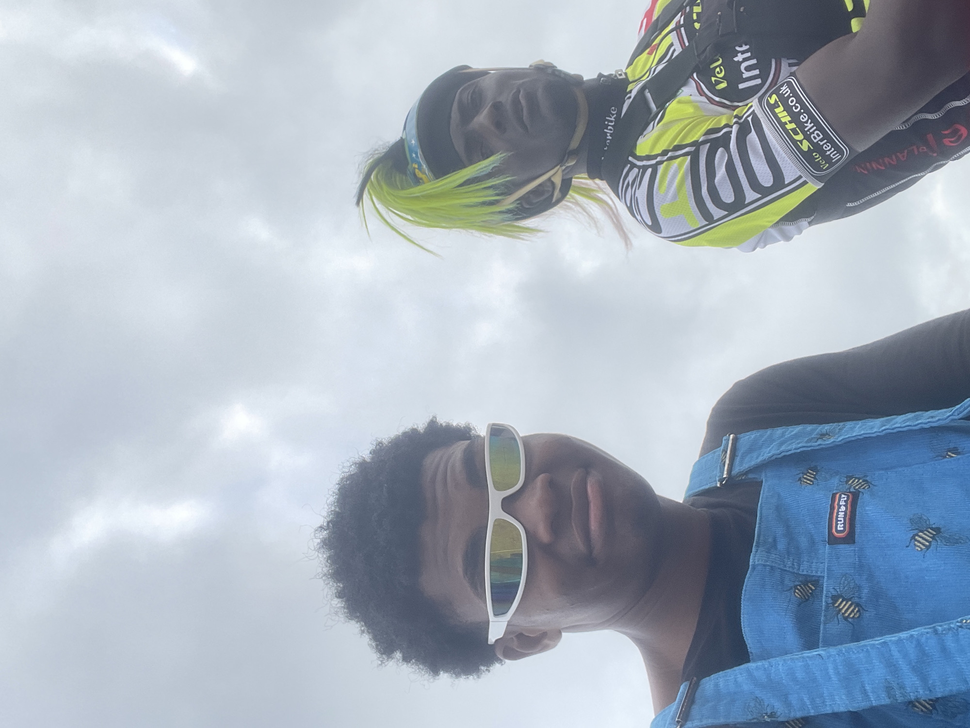 The image shows two young Black individuals against a cloudy sky, with one person on the left wearing a black t-shirt and bright blue overalls adorned with a bee pattern, and white wrap-around sunglasses. The person on the right is wearing a black cycling helmet with neon green hair extensions and a brightly colored cycling jersey with various logos, including "Velo Schils InterBike.co.uk". The sky is overcast and appears to be a dull gray with some lighter patches indicating breaks in the clouds. The individuals appear to be outdoors, possibly in a park or open area, given the sky and lack of urban elements. The location is specified as Inupa Kekere, Nigeria. No specific activities or interactions between the individuals are discernible from the image, though their attire suggests participation in outdoor activities like cycling or a casual outing.