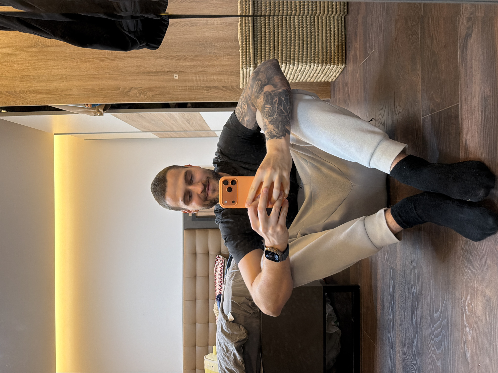 A man is taking a selfie in a mirror. He is sitting on a wooden floor, wearing black socks, light-colored pants, and a black t-shirt. He has a watch on his left wrist and tattoos on his left arm. He is holding an orange phone. In the background, a bed with a headboard and bedding, a nightstand with an object on it, a wardrobe, and a woven basket can be seen. The image is possibly taken in a residential setting. Location details regarding the city of Sofia, Bulgaria are not discernible from the image.