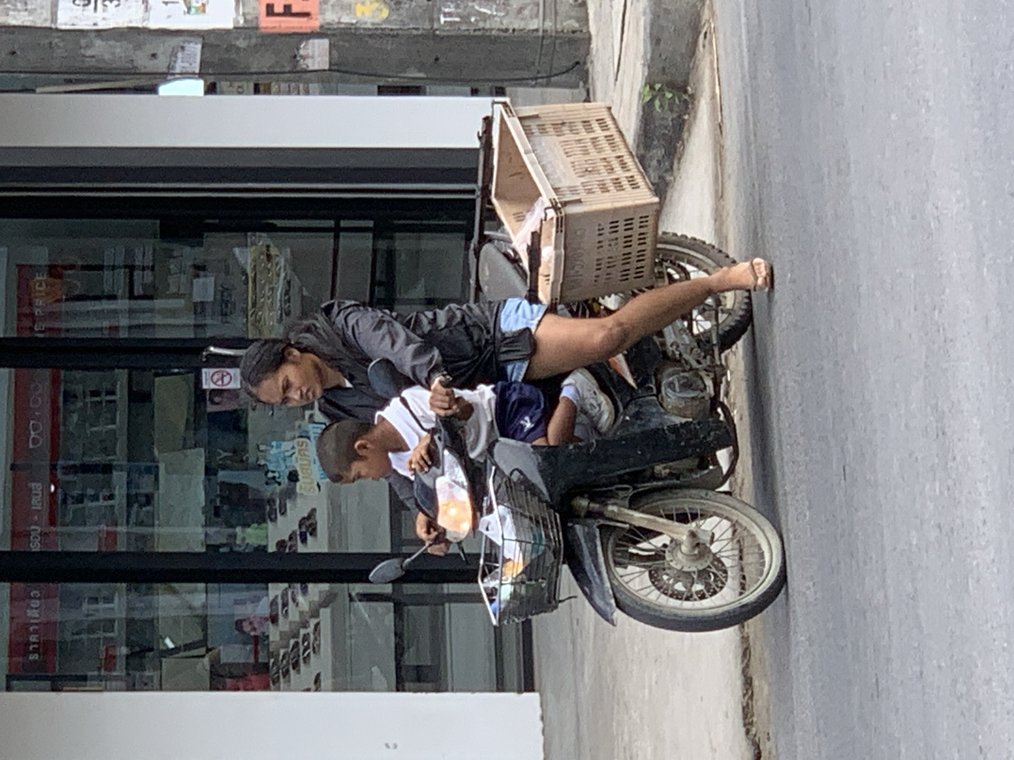 An adult female wearing a black hooded jacket and light blue shorts is operating a motorcycle. A young boy, wearing a white t-shirt and dark blue shorts, is seated in front of her on the motorcycle. Both individuals are facing forward, appearing to be in motion. The motorcycle has a basket attached to the front and a larger, light brown crate secured to the rear. The background shows the exterior of a building with large glass windows, displaying various items. The scene is set on a paved road in front of a sidewalk. The overall impression is a snapshot of daily life in a commercial area, likely in the city of Ko Samui District, Thailand, given the visible Thai script on signage.