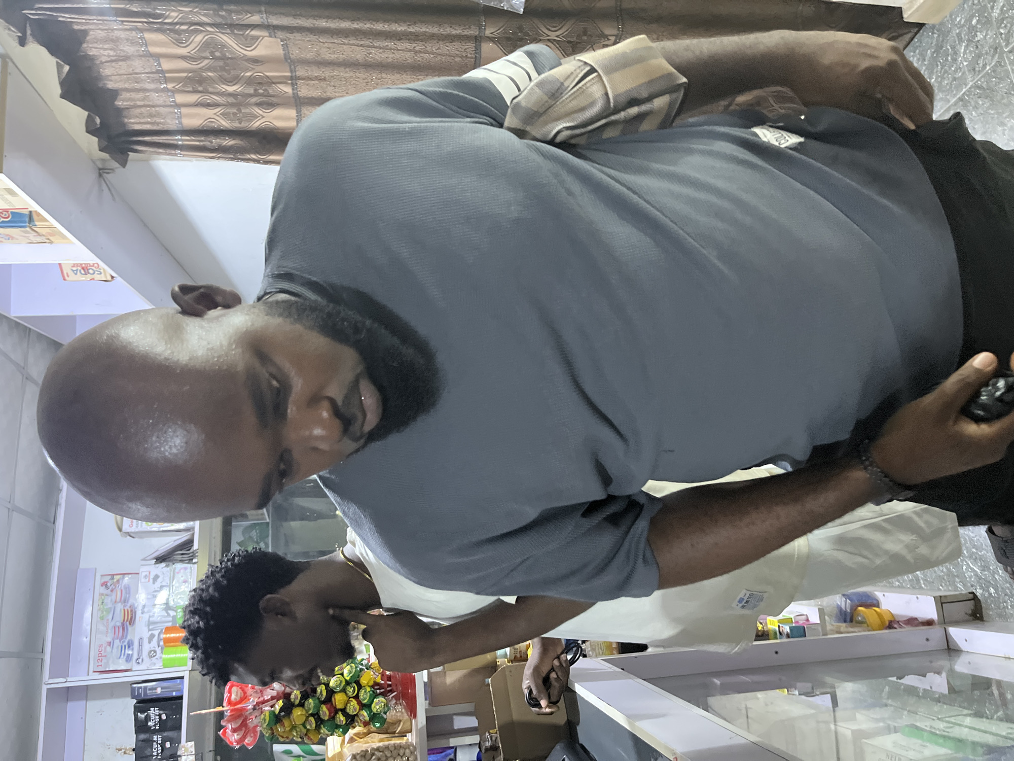 A bald man with a beard is standing in what appears to be a store. He is wearing a grey shirt and black pants, holding a black object in his right hand. Behind him, another person is visible, also appearing to be in the store. Shelves stocked with various items are evident in the background, along with decorations. The location is likely in Dushepe, Nigeria.