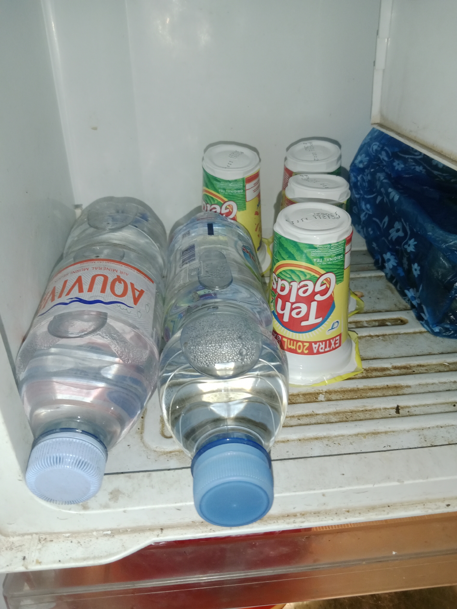 The image shows the interior of a refrigerator. Two large plastic bottles of Aquavita mineral water are positioned on the middle shelf. To the right of the water bottles are several small cans of Teh Gelas, a brand of packaged tea. One can of Teh Gelas is clearly visible, with its yellow and green labeling depicting a tea leaf and the text "Teh Gelas" and "EXTRA 20ML". Behind the Teh Gelas cans, a blue plastic bag with a floral pattern is visible. The refrigerator shelves appear to be made of white plastic and show some signs of dirt and grime. The overall environment is a domestic refrigerator in what could be Ringinrejo, Indonesia, given the brands of beverages present.