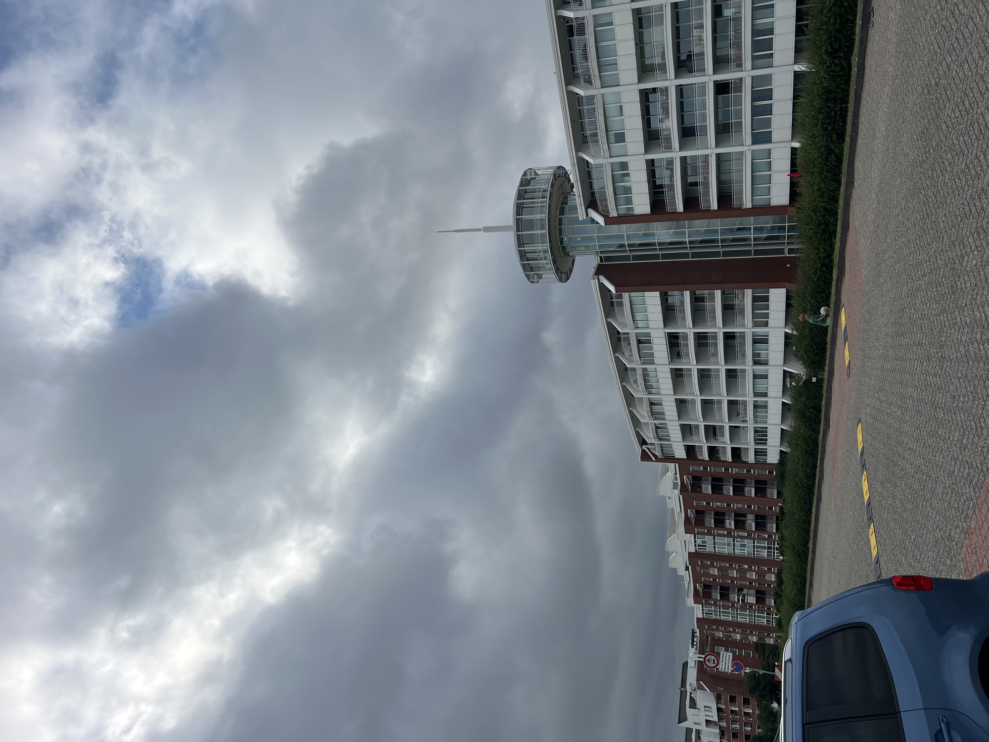 A portrait-oriented, downward-angled shot shows a modern building complex in Wilhelmshaven, Germany, under a heavily clouded sky. The building on the right is white with multiple balconies and glass-fronted windows. A distinctive circular structure with a spire juts out from a connecting section. Below it, a series of red brick buildings with white-trimmed windows continues the architectural line. In the foreground, a blue car is parked on a grey, patterned paved surface. A low green hedge runs along the base of the buildings, separating them from the pavement. Two figures are visible near the hedge: one in a green shirt appears to be interacting with a white object, possibly a ball, while another individual in a red garment stands further back. The weather is overcast, with dramatic, dark grey clouds dominating the sky, interspersed with lighter, brighter patches, suggesting a potentially stormy or unsettled atmosphere. The time of day is not explicitly discernible due to the weather conditions, but the general lighting suggests daytime. The scene is one of a modern urban or residential environment, with a focus on the contemporary architecture and the interplay of clouds and structures.