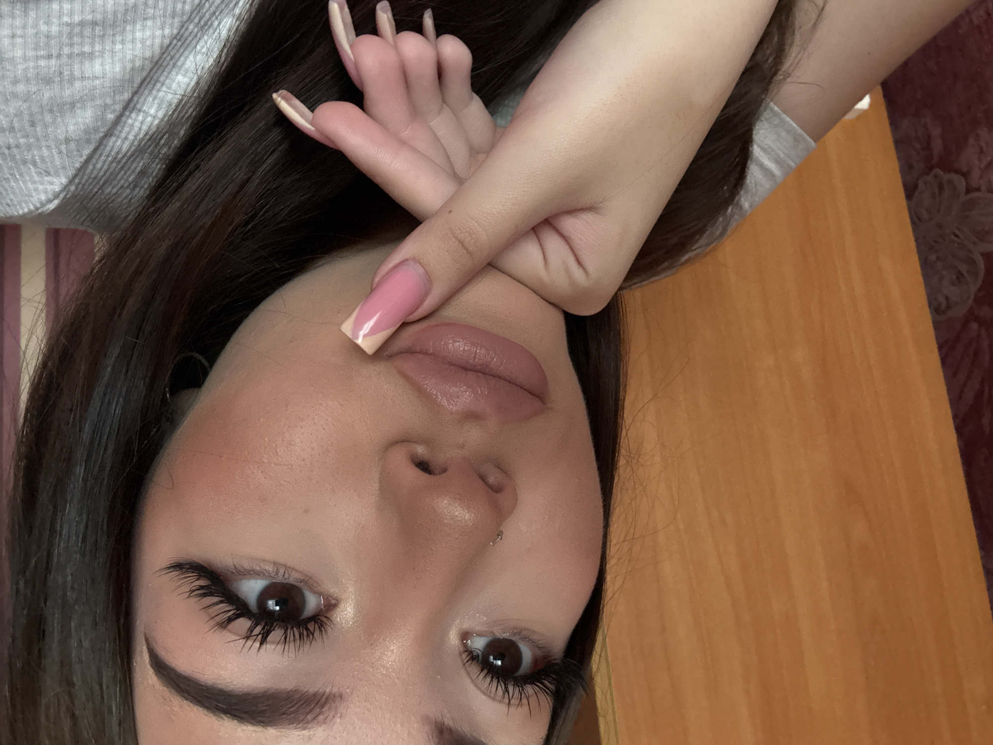 A close-up, downward-facing image shows a woman with dark brown hair, light skin, and brown eyes. She is wearing heavy mascara on her upper and lower lashes. Her lips are light pink and glossy. She has a silver nose piercing in her left nostril. Her left hand is placed across her face, displaying long, square-shaped nails with a pink and beige color scheme. She appears to be wearing a grey ribbed top. A wooden surface is visible on the right side of the image. Behind her are a wall and a textured, floral-patterned fabric. The image does not contain sufficient information to determine its location in Belopakhotnyi, Russia.