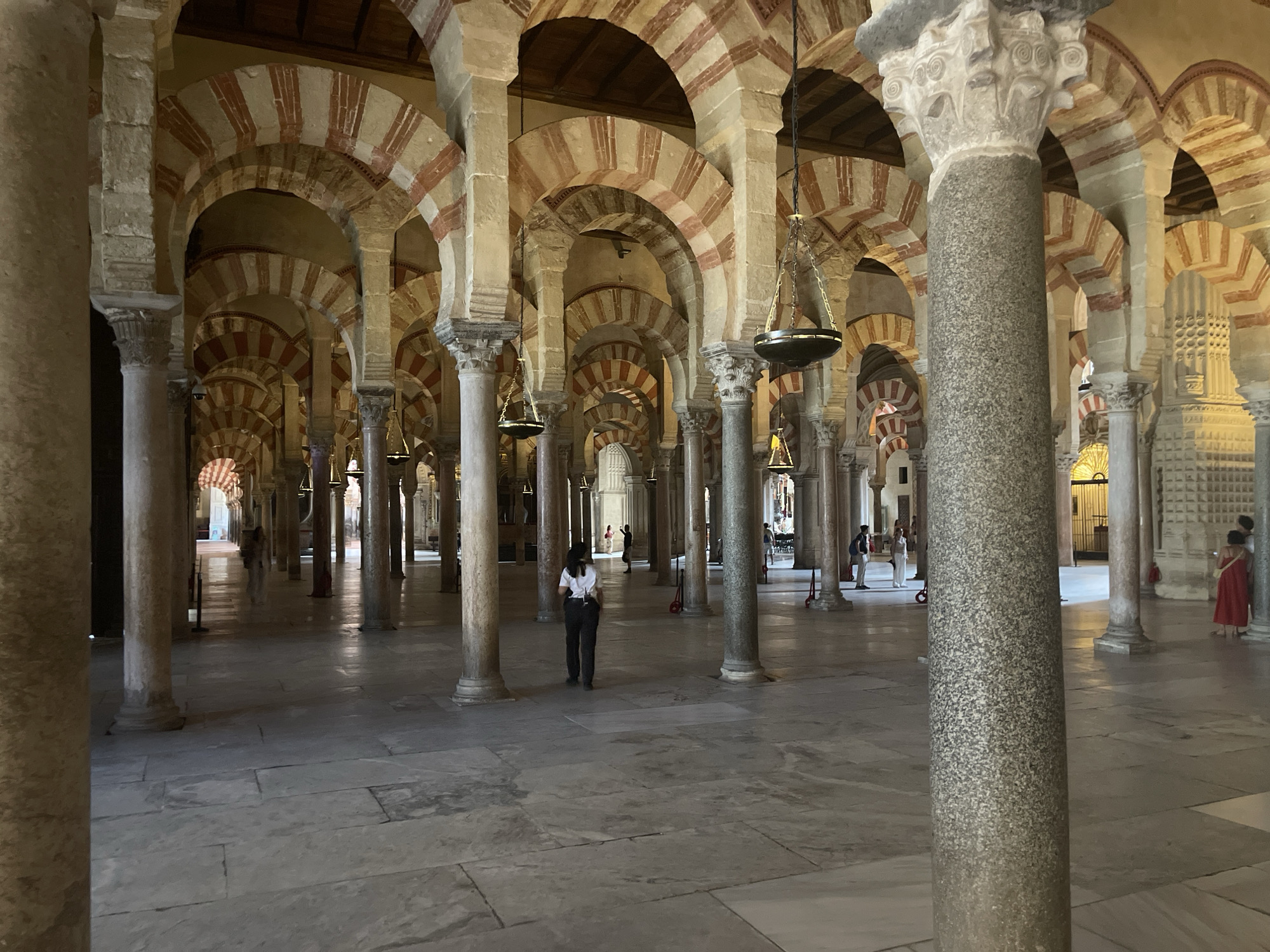 The image depicts the interior of the Great Mosque of Córdoba, Spain, an iconic architectural landmark. The scene is dominated by the characteristic horseshoe arches with red and white voussoirs supported by numerous columns. The floor is paved with large, light-colored stone tiles, reflecting the light from above. Several people are visible throughout the hall, appearing as tourists or visitors. In the foreground, a person is walking away from the camera, wearing a white shirt and dark trousers. Further in the background, other individuals can be seen in small groups or standing, observing the architecture. Hanging from the ceiling are ornate, dark-colored lanterns. The vastness of the space is emphasized by the repeating rows of arches and columns extending into the distance.