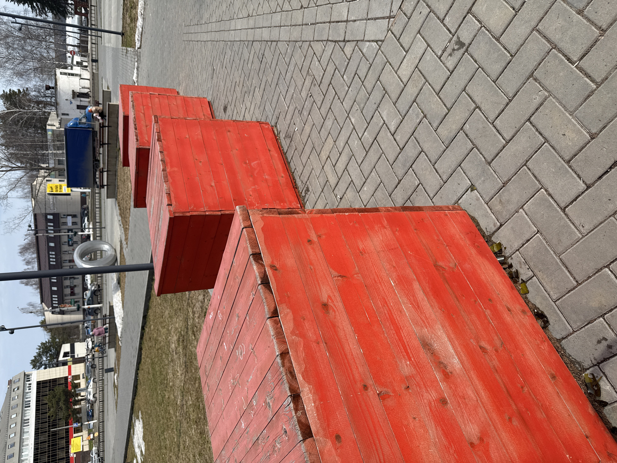 A ground-level perspective shows four progressively smaller red wooden boxes arranged in a diagonal line on a gray herringbone-patterned brick surface, suggesting a city setting. In the background, partially obscured by bare trees, are buildings, parked vehicles, and streetlights. A grassy area with small patches of snow is visible on the left side of the boxes.