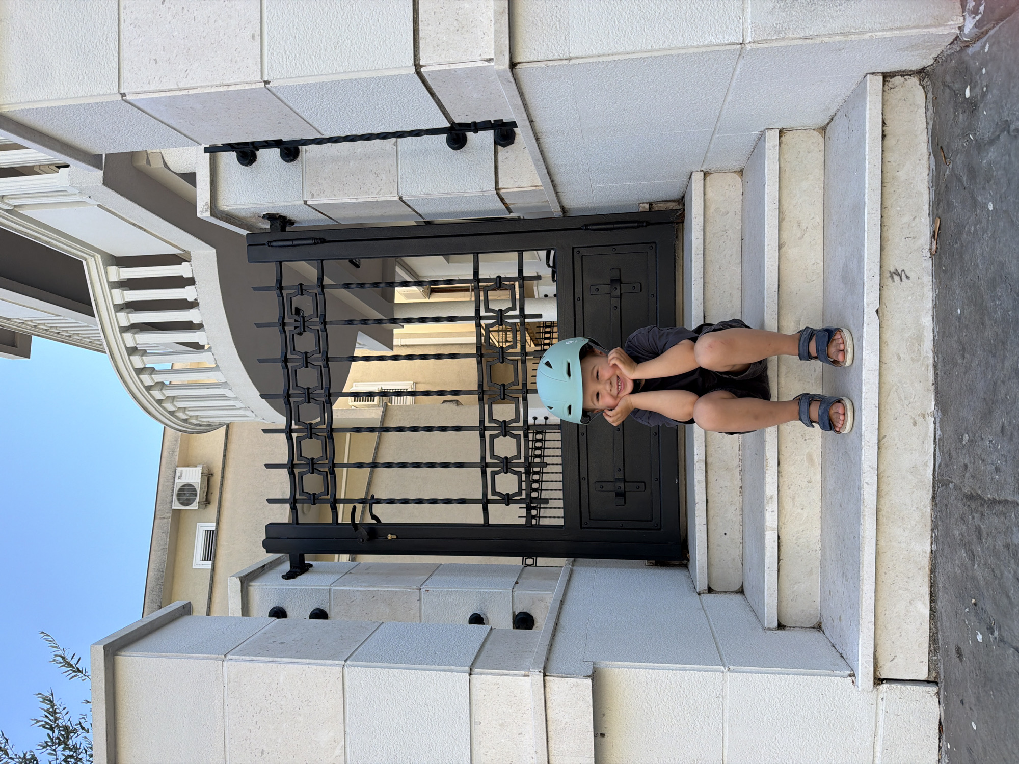 A young child wearing a light blue helmet sits on white stone steps in front of a black metal gate, smiling and holding their hands to their face. The steps lead up to the gate which is set into a wall with beige stone accents. A building with a curved balcony is visible to the left of the gate, with an air conditioning unit mounted on the wall. The sky is visible in the upper left corner. The setting is likely Budva, Montenegro, based on architectural characteristics.