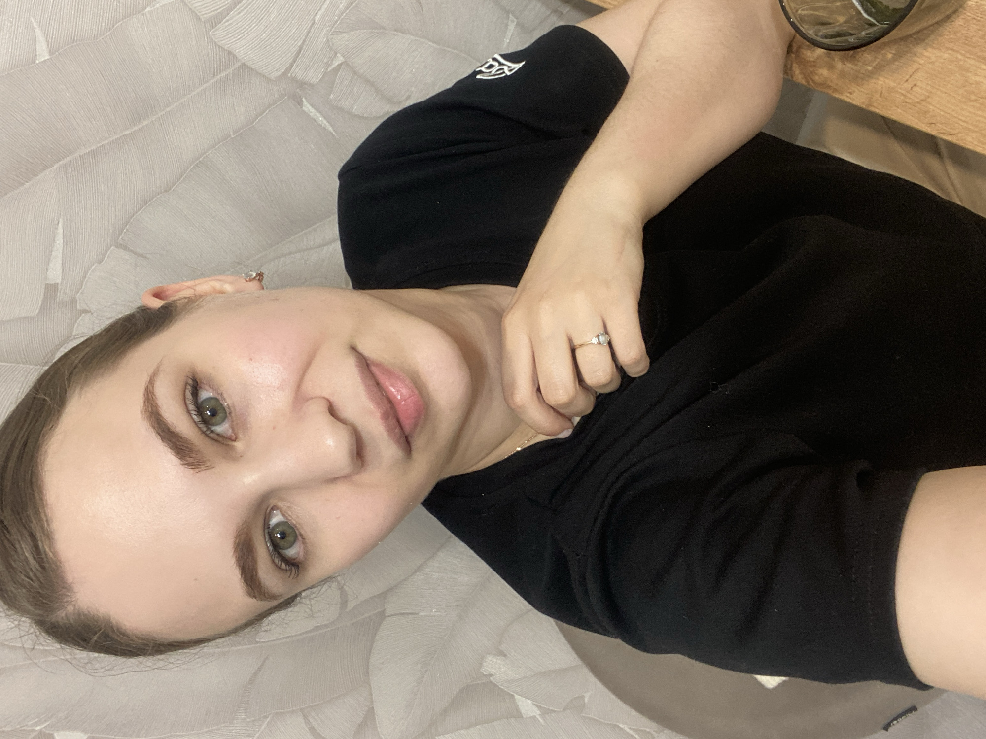 The image presents a close-up, head-and-shoulders portrait of an adult female, captured indoors. She has light skin, green eyes, and light brown hair pulled back from her face. Her expression is neutral with a slight smile, as she looks directly into the camera. She is wearing a black, short-sleeved t-shirt, which features a white emblem on the right sleeve. A thin gold chain necklace is visible around her neck, and she wears small, gold-toned hoop earrings. On the ring finger of her left hand, a gold ring set with a single clear stone is present. Her left hand is raised to her upper chest area, with fingers slightly curled and resting against her t-shirt.
The immediate background consists of a light-colored wall with an abstract, textured pattern resembling overlapping leaves or layered brushstrokes. To the left, a segment of a dark, possibly upholstered, furniture item is partially visible. In the bottom right corner of the frame, a portion of a clear glass or cup, containing visible liquid or ice, rests on a light-colored wooden surface. The specific location within the city of Chaikovsky, Russia, cannot be determined from the visual information provided in the image.