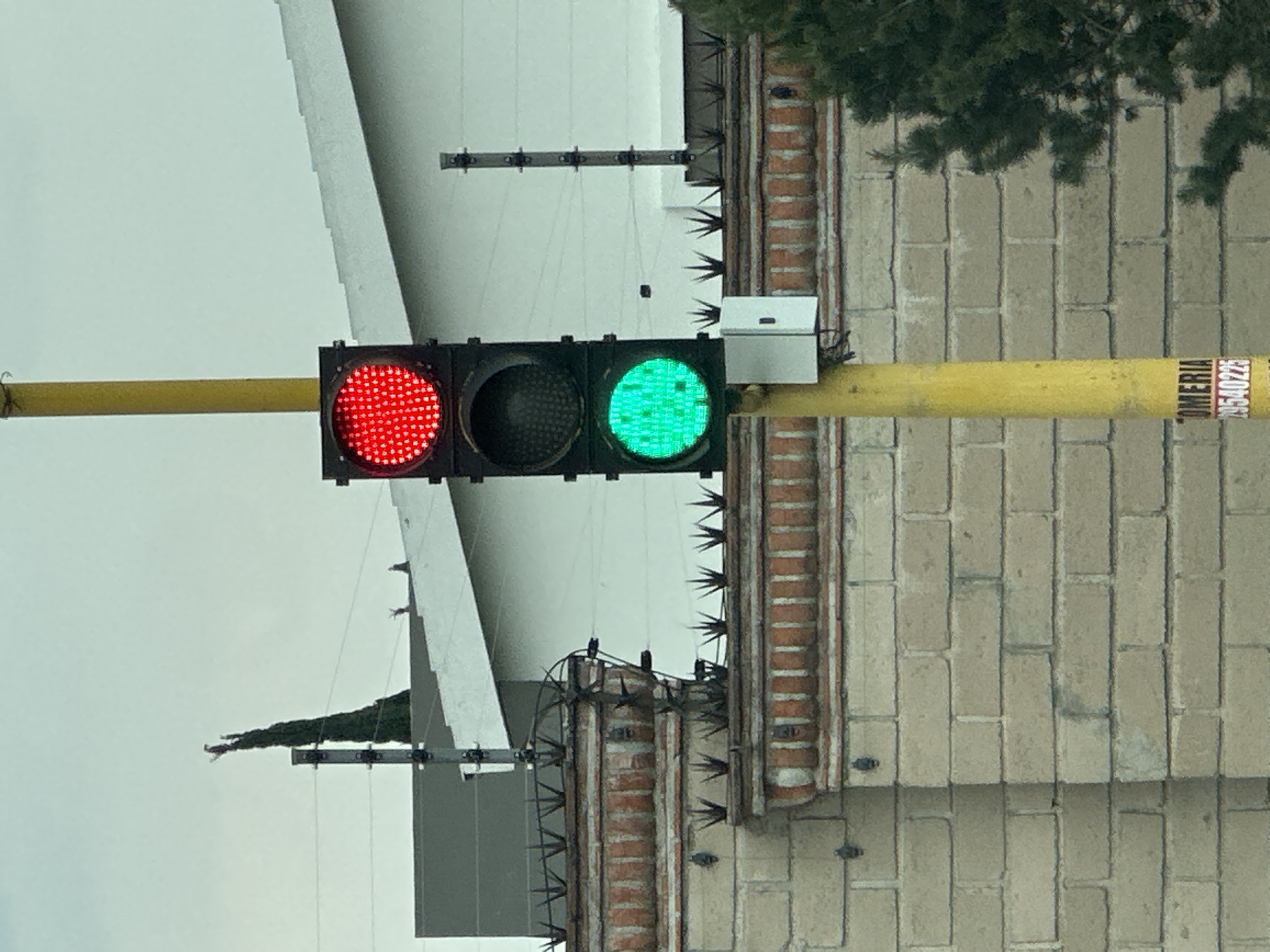 A street-level, medium-shot photograph depicts a traffic light in San Bernardino Tlaxcalancingo, Mexico. The signal shows a red light illuminated to the left and a green light illuminated to the right, with the central light unlit. The light fixture is affixed to a horizontal yellow pole. Above and behind the light, architectural features are visible, including a white building section, a brick structure with anti-climbing spikes, and foliage. A partial sign is visible on the right. The sky is overcast.
