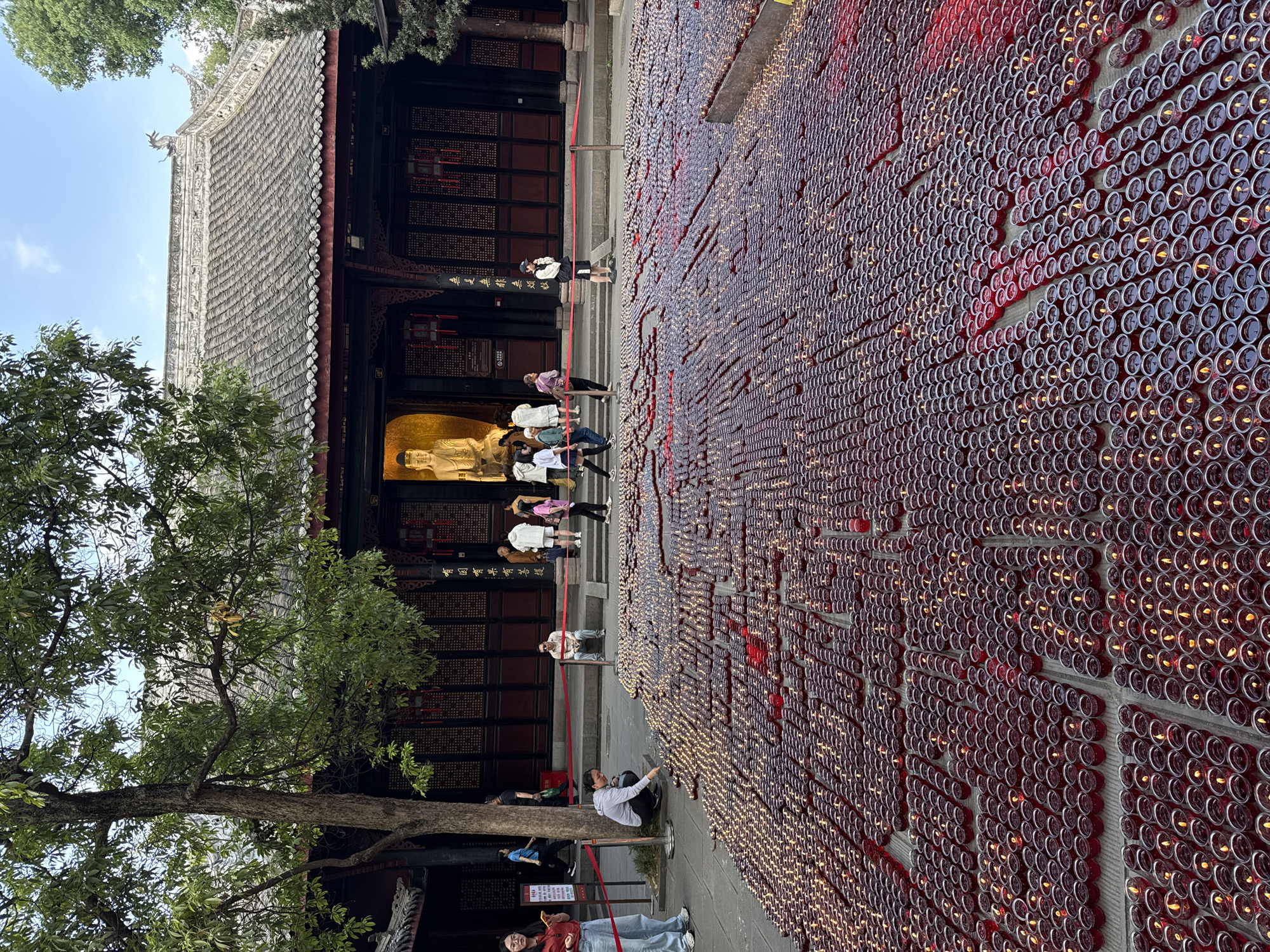The image shows a courtyard in front of a traditional Chinese building. The courtyard is filled with rows upon rows of small, round candle holders, likely made of glass or metal, with lit candles inside, creating a warm, glowing effect. A group of people are ascending the steps of the building, while others are standing or sitting nearby. A tree stands to the left of the building, its branches partially obscuring the view. The building has a traditional Chinese architectural style with a tiled roof and intricate wooden details. Based on the visual evidence, including the architectural style and cultural elements, it is likely located in the Qingyang District, China. The scene captures a moment of religious observance or memorial.