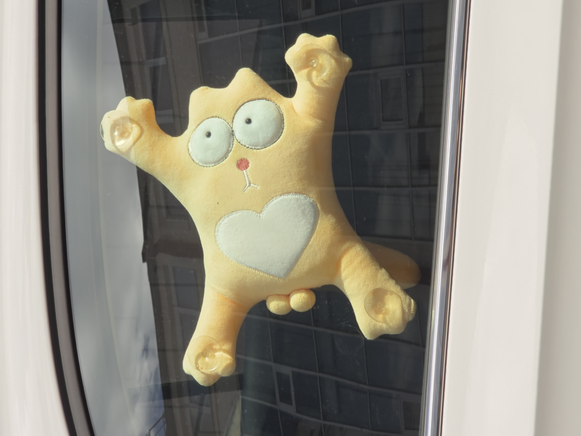The image shows a yellow plush toy, resembling a cartoon character, attached to the inside of a car window with suction cups. The toy has large, round, white eyes with black pupils, a small red nose, and a white heart shape on its chest. The car is cream-colored. Reflections on the car window show a building with multiple floors and windows. The image is a close-up shot, focusing on the plush toy and the car window. The provided metadata states the image was taken in Rostov-on-Don, Russia, on July 1, 2024, at 3:04 PM.