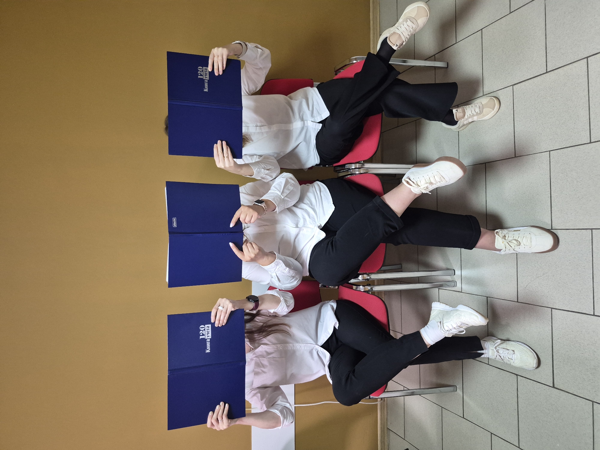 Three people sit in red chairs, each holding a blue booklet in front of their face. They wear white shirts and black pants. Two have light-colored sneakers and one has white sneakers. The setting appears to be indoors with a beige wall and tiled floor. The text on the booklet reads "120 Kniga ycheta". The picture was taken in the city of Chaikovsky, Russia.