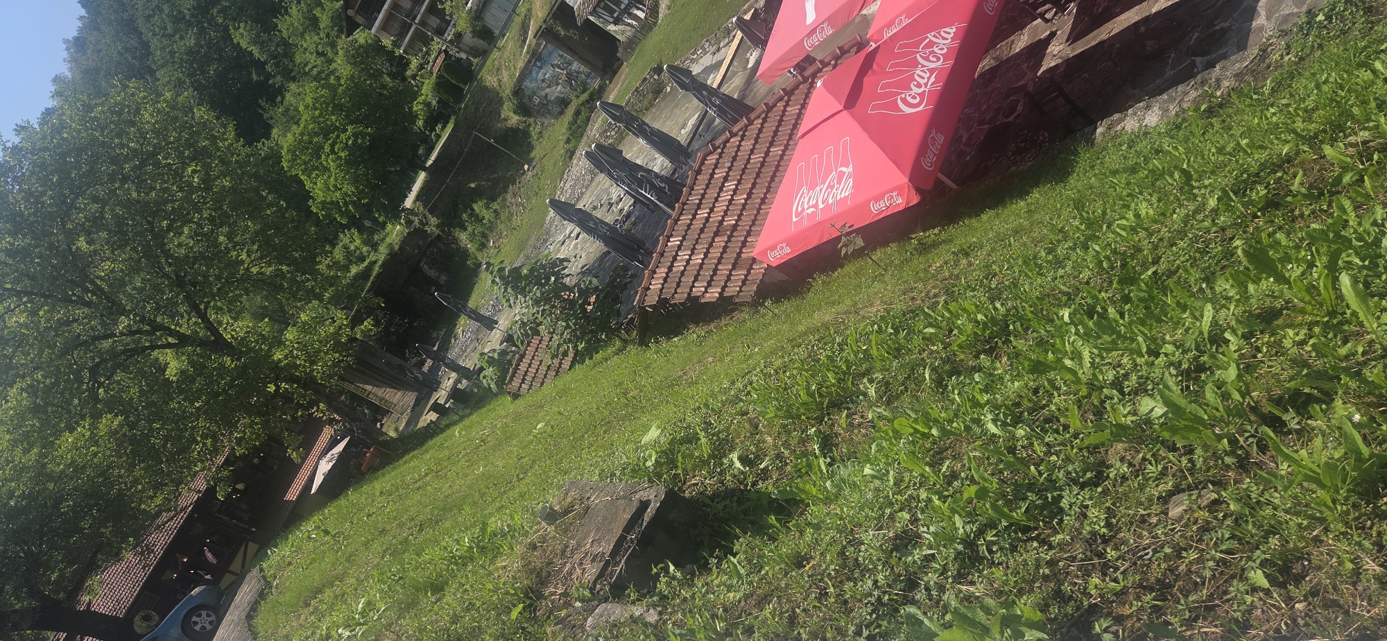 The image, taken with a tilted perspective, depicts an outdoor scene in Beli Osam, Bulgaria, under clear, sunny conditions. In the lower right, a series of red umbrellas with the Coca-Cola logo are arranged, indicating an outdoor seating area. Adjacent to these umbrellas is a building section with a reddish-brown tiled roof. To the left of the umbrellas, a steep, grassy embankment dominates the lower foreground, showing varied green vegetation.
Further into the scene, behind the initial tiled roof, is a flowing body of water, likely a river or stream. Along its banks, additional outdoor umbrellas, appearing dark in color, are visible. On the opposite side of the water, a paved road runs, and beyond it, a structure with a painted mural is discernible on a retaining wall. Another multi-story building with balconies is situated further up on the far bank.
In the upper left portion of the image, a light blue car is parked on a surface that appears to be part of a wooden deck or paved area, adjacent to a larger building. This building features a reddish-brown tiled roof, similar to the one in the foreground, and light-colored walls. Abundant green foliage, consisting of numerous trees with dense canopies, frames the buildings and lines the surrounding hillsides. The background is dominated by heavily forested hills rising under a clear blue sky. No explicit human figures or direct interactions are clearly visible.