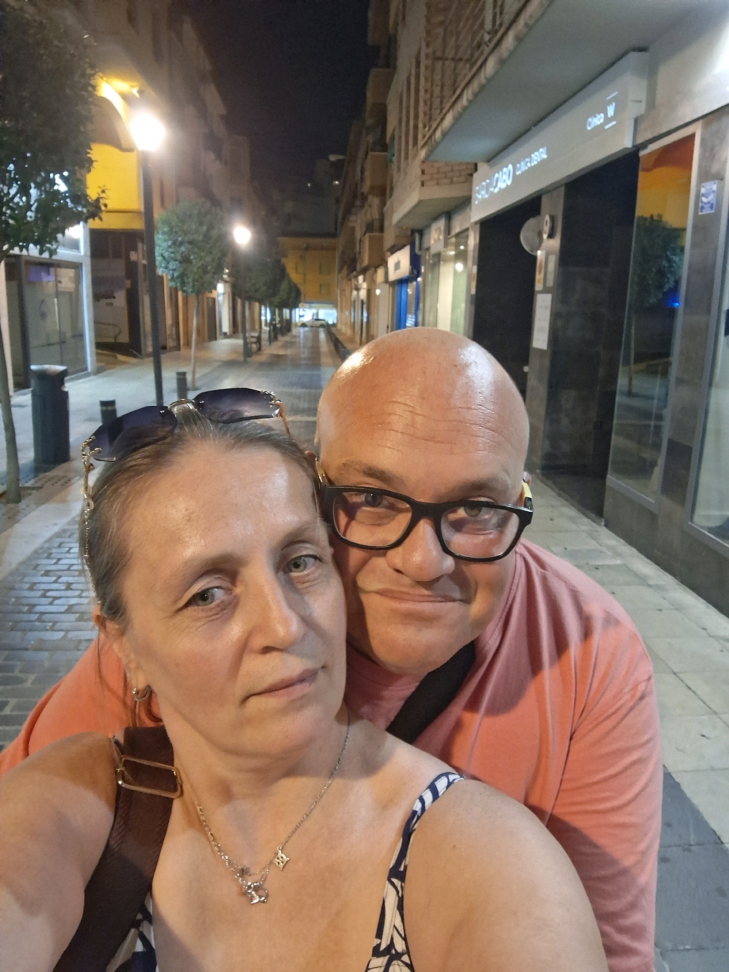 This is a selfie taken at night on a street in Alcañiz, Spain. A bald man wearing glasses and a pink t-shirt is in the foreground, partially obscuring the view of a woman next to him. The woman has sunglasses perched on her head and is wearing a necklace. They are standing on a cobblestone sidewalk lined with trees and streetlights. In the background, buildings with illuminated signs, including one that reads "SARO-CABO CLINICA DENTAL," are visible. The street appears wet, suggesting recent rain.