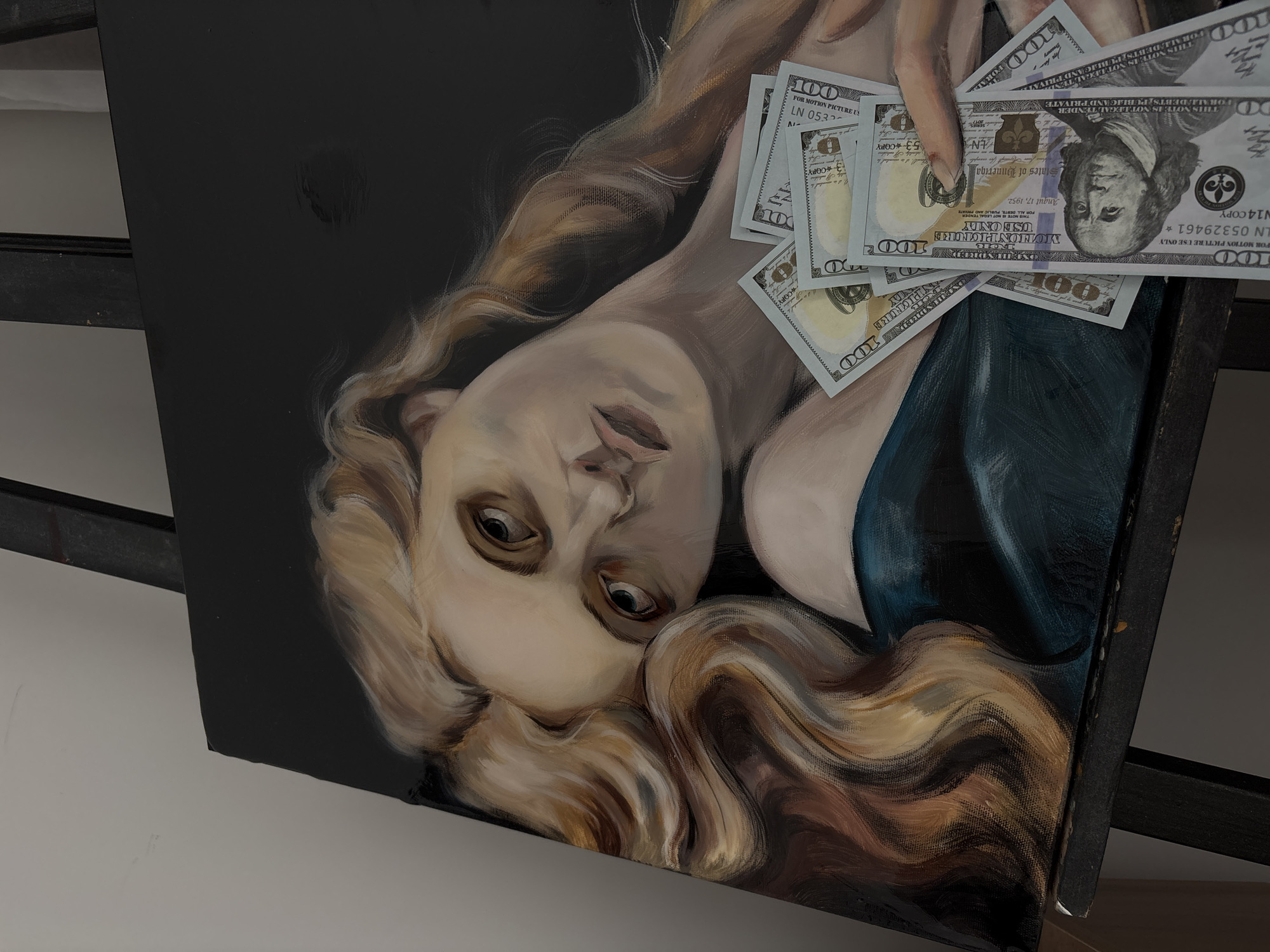 The image is a photograph of an oil painting. The painting depicts a pale-skinned woman with long, flowing blonde hair, lying down. Several 100 dollar bills are held in a hand above her. The painting is set against a dark, nearly black background. It is on an easel.