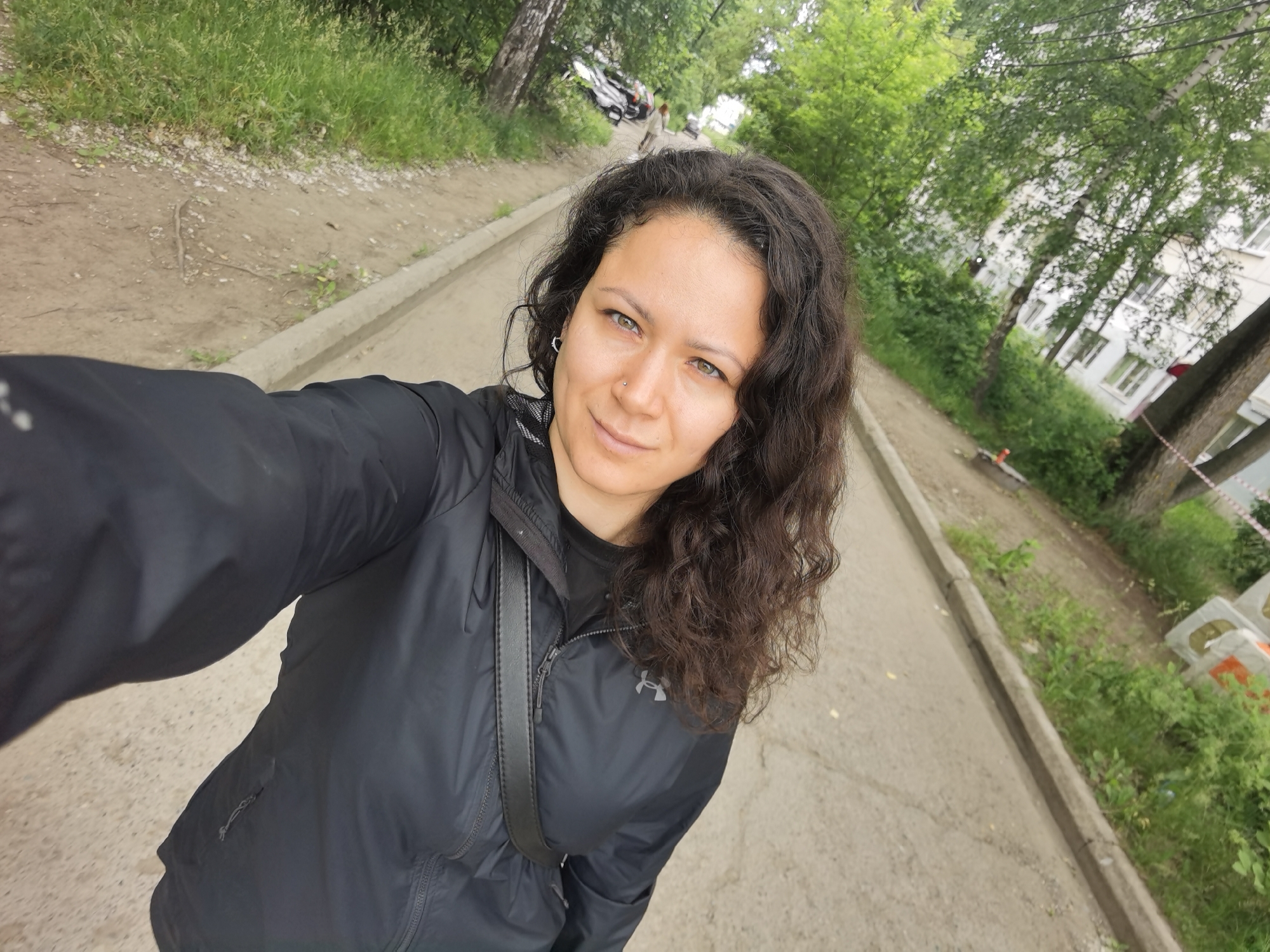 A woman with curly dark brown hair is taking a selfie outdoors. She is wearing a black jacket with a zipper and a black strap across her chest. She has a nose ring and an earring. The background shows a pathway, trees, and a building. The scene appears to be a residential area in Chaikovsky, Russia, possibly near a park or a walkway.