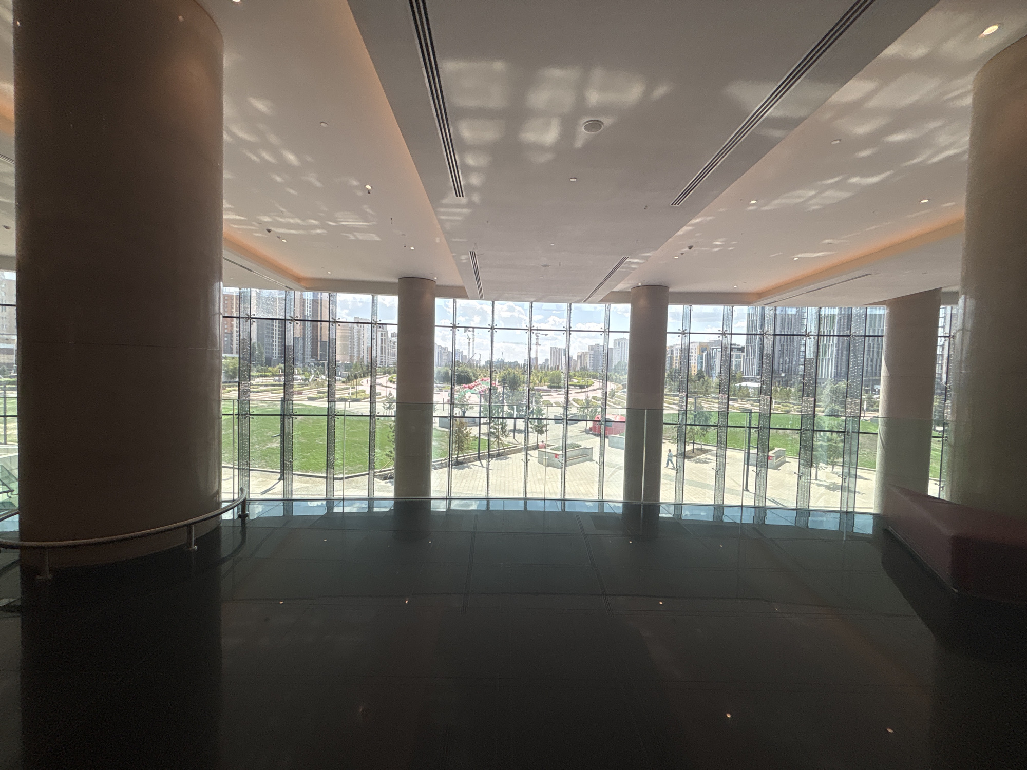 The image depicts the interior of a modern building, likely a convention center or office building, with a large, polished black floor reflecting the light. Two substantial, cylindrical columns are present, one on the left and another on the right, supporting the building's structure. A panoramic window wall dominates the background, offering a view of an urban landscape.
The exterior scene shows a well-maintained green park with manicured lawns and scattered trees. Beyond the park, a cityscape unfolds with numerous contemporary buildings, many appearing to be residential high-rises. Several construction cranes are visible, indicating ongoing development in the area. The sky is bright with scattered clouds. A person is walking in the park area visible through the window.
The architectural style and the presence of the modern buildings and infrastructure strongly suggest the location is Astana, Kazakhstan. The overall impression is one of a new and developing urban environment characterized by modern architecture and expansive green spaces.
