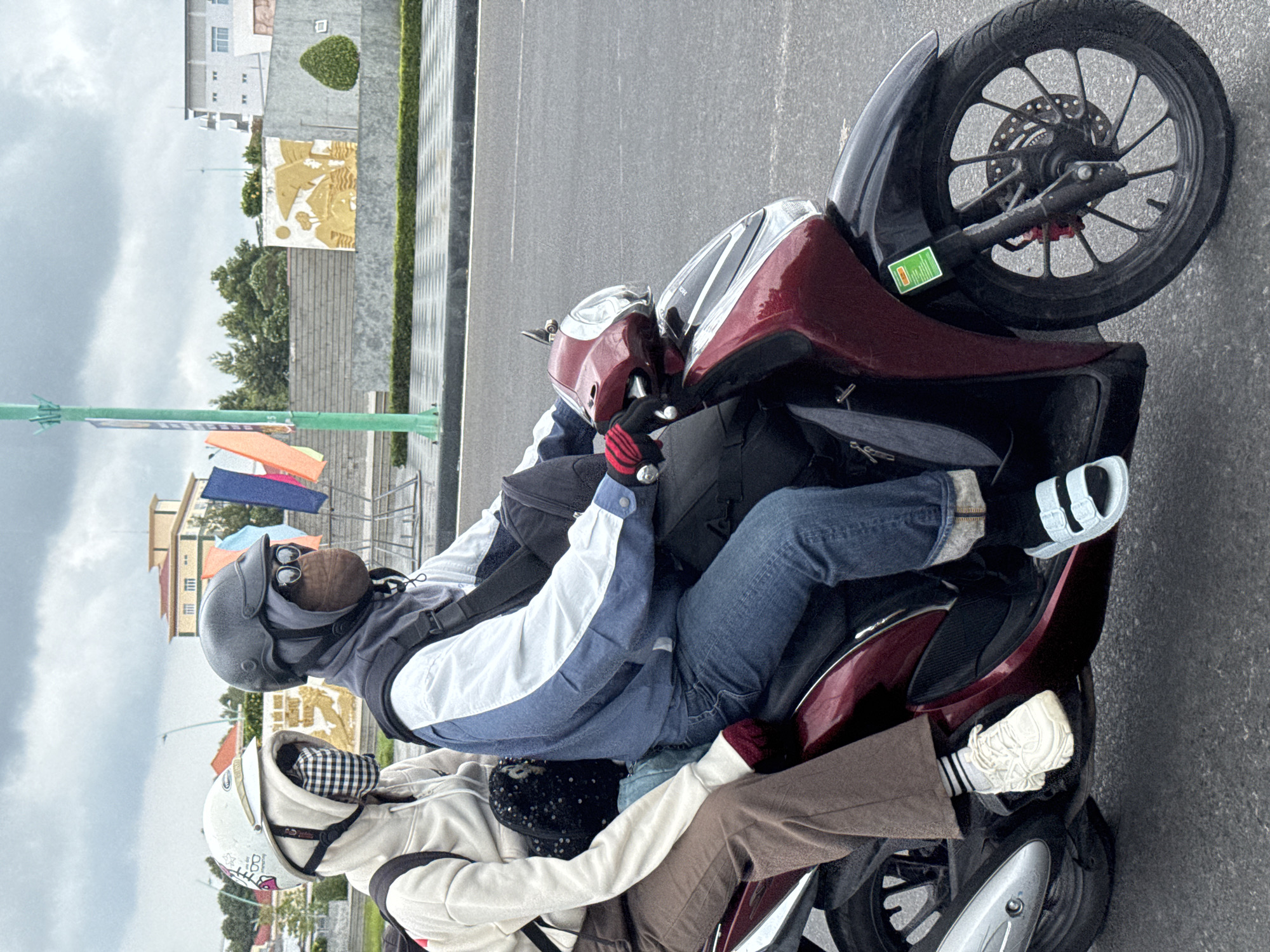 Two individuals are riding on a maroon and black scooter on a paved road in Phan Thiết, Vietnam. The front passenger, wearing a white helmet and a beige hooded jacket, has their face covered with a black and white checkered cloth. They are also wearing brown trousers and white athletic shoes. The driver, wearing a dark helmet, sunglasses, and a grey balaclava, has their face partially covered with a brown cloth. They are wearing a blue and white jacket, blue denim jeans, and light blue slide sandals. Both individuals are wearing gloves. The scooter has a green sticker on its front. In the background, there is a pedestrian walkway, a wall with a gold mural depicting a landscape, and several buildings. The sky is overcast.