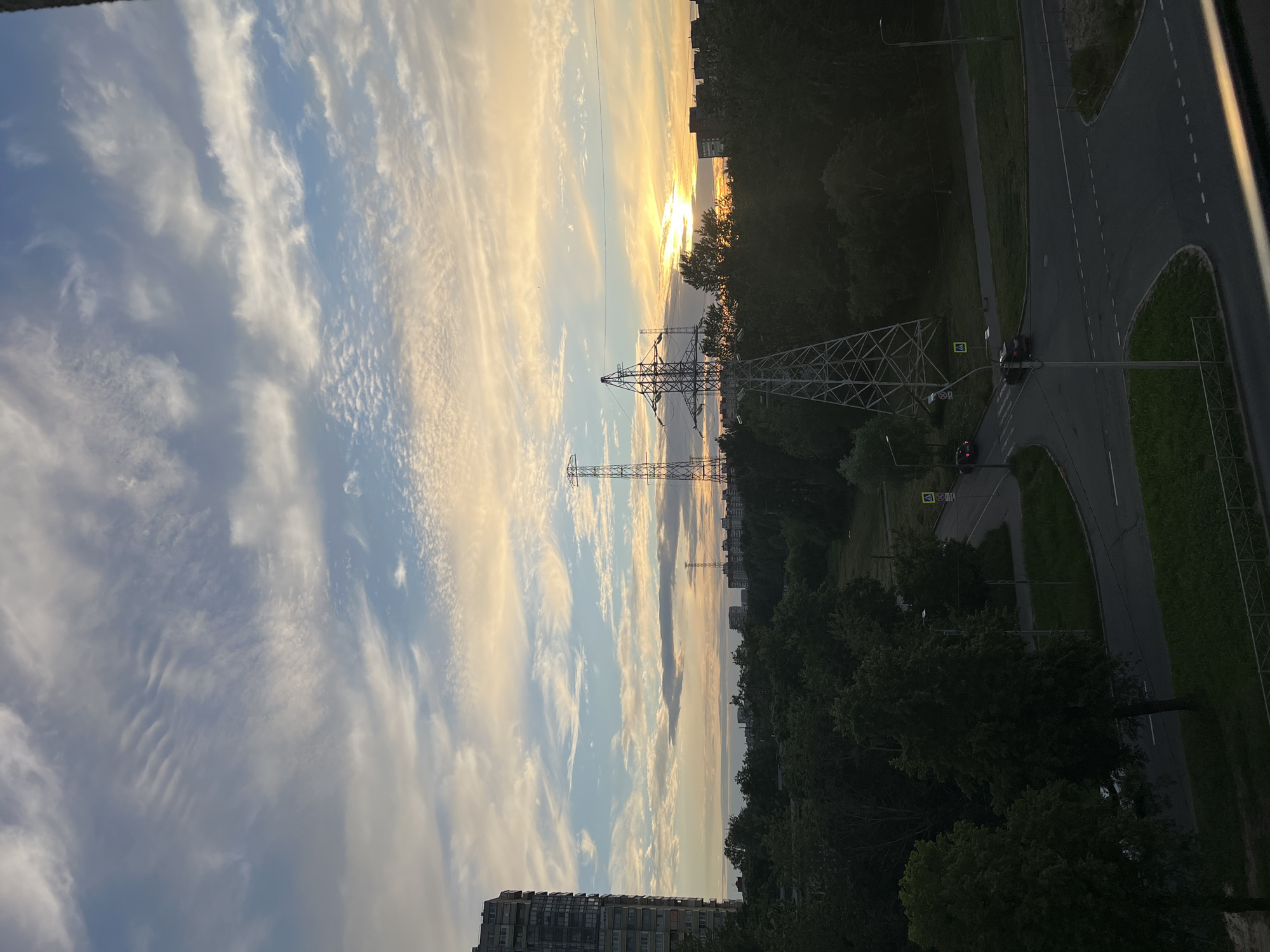 The image captures a view of the sky over a city park or suburban area during a sunset. In the foreground, on the left, is a tall residential building with numerous balconies. To the right of the building, several power line towers are visible, silhouetted against the sky, with the setting sun peeking through the trees and clouds on the horizon. The sky is filled with a dramatic display of clouds, ranging from wispy cirrus formations to more textured, mottled clouds, colored with shades of orange, yellow, pink, and blue from the sunset. Below the sky, a dense canopy of green trees stretches across the middle ground, obscuring much of the ground. A road network with cars and streetlights is visible within the tree line and extending into the foreground. The scene appears to be an urban or semi-urban environment in Saint Petersburg, Russia, characterized by a mix of modern architecture, industrial infrastructure (power lines), and green spaces.
