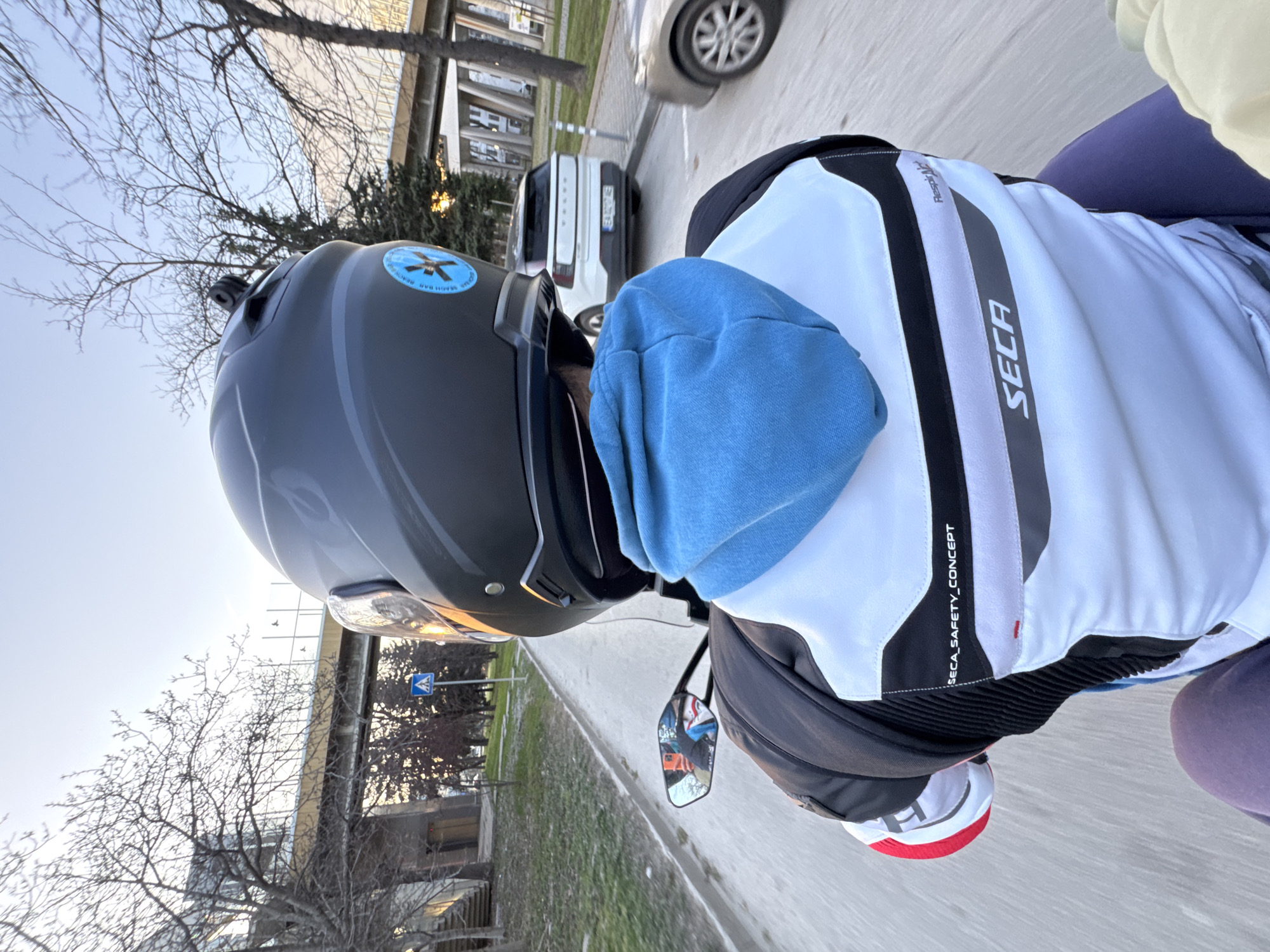 A person is riding a motorcycle or scooter on a road in Sofia, Bulgaria. The rider is wearing a black helmet with a sticker on the left side, a blue hooded sweatshirt, and a white SECA jacket with black and gray accents. The rider is facing away from the camera. The motorcycle's rearview mirror is visible. The background includes trees without leaves, buildings, and a white car. A pedestrian crossing sign is visible. The sky is bright and partially cloudy.