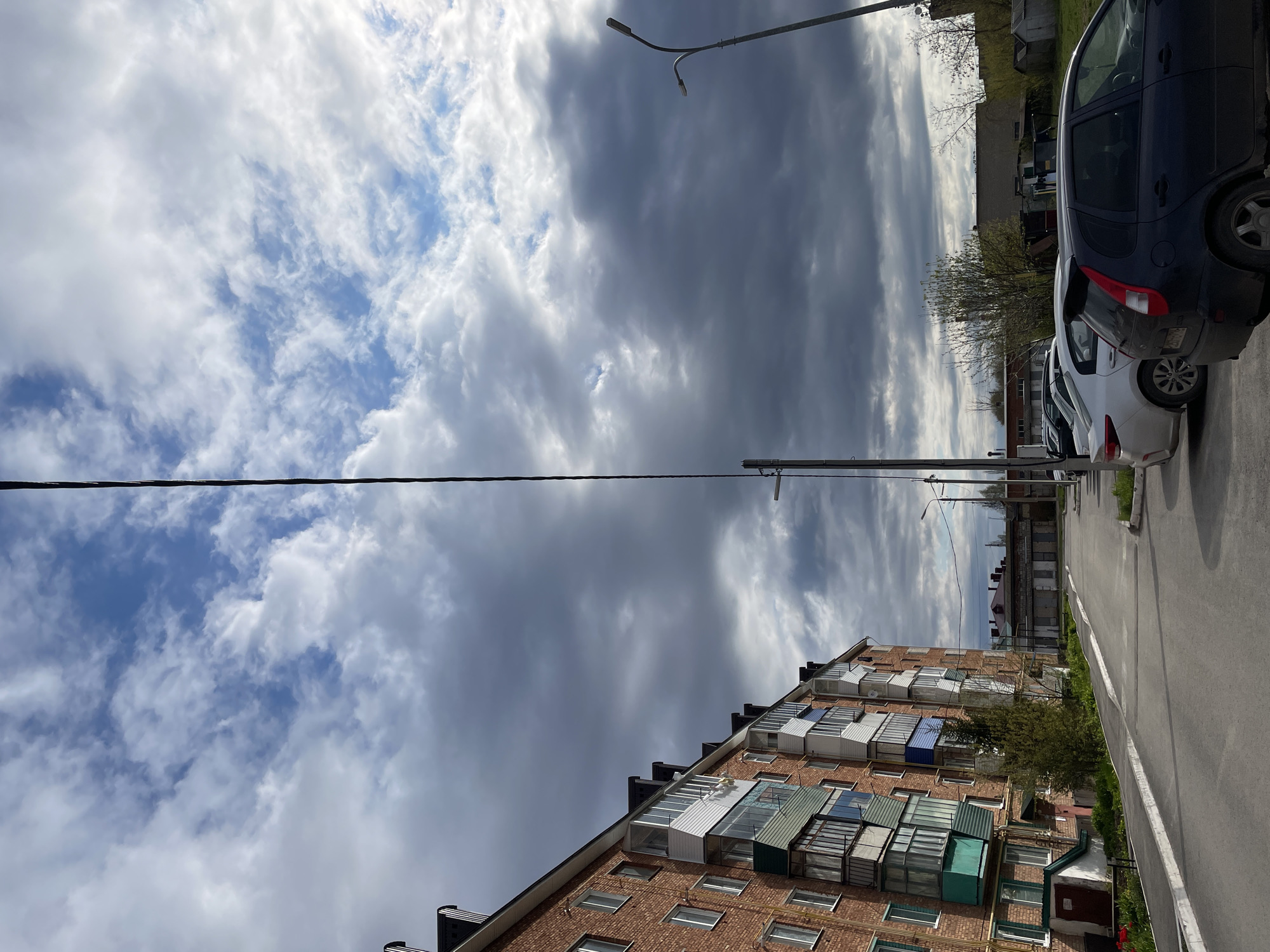 The image shows a street in Urussu, Russia, with an apartment building on the left, cars parked on the right, and a cloudy sky overhead. The apartment building is made of red brick and has multiple balconies of various colors and designs. A utility pole with a power line spans across the top half of the image. A street lamp is visible above the parked cars. The road appears to be asphalt. No people are visible in the shot. The weather is overcast.