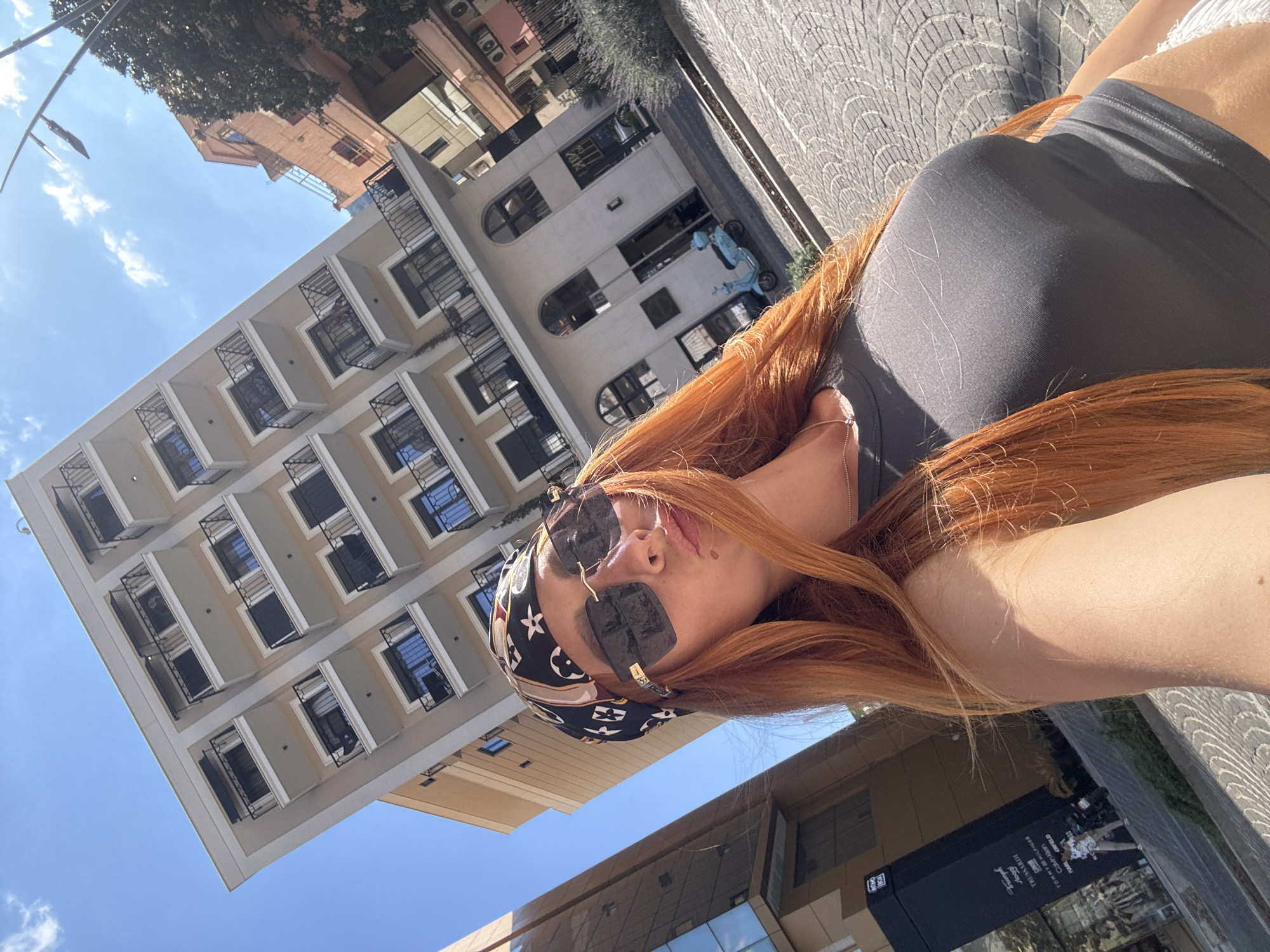 The image is a selfie taken by a woman with long, auburn hair, wearing a black bandana with a repeating pattern of logos, oversized dark sunglasses, and a dark gray ribbed tank top. She is posing with her lips pursed and her head tilted. The background shows a city street scene with modern buildings under a clear blue sky with scattered clouds. To the left, a tall building with glass facades is visible, featuring multiple floors with reflective windows and a black storefront at its base. To the right, a multi-story building with balconies and beige-colored cladding dominates the upper portion of the image, with an orange building peeking out behind it. A light blue scooter is parked on the sidewalk in front of a building with arched windows. The street is paved with cobblestones. The location is identified as the city of Budva, Montenegro.