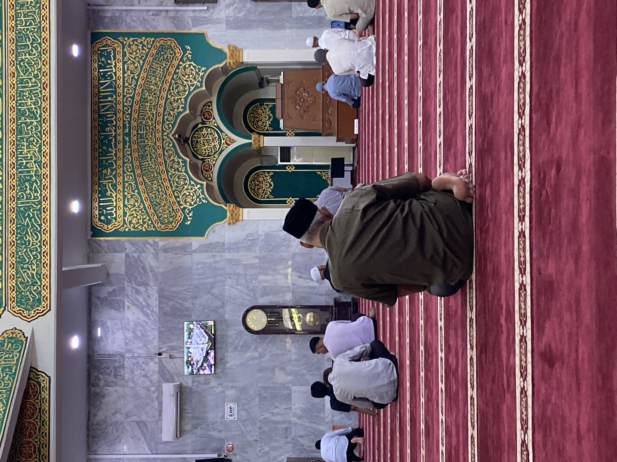 The image depicts the interior of a mosque, likely in Lhokseumawe, Indonesia, with a focus on a group of men engaged in prayer. The floor is covered with a deep red carpet featuring parallel white and gold striped patterns, indicating designated prayer lines. In the foreground, a man wearing a dark olive green shirt and black cap is seated on the carpet, facing away from the camera. His legs are bent, and his bare feet are visible.
Behind him, several other men are also seated or kneeling in prayer. To the left, two men in white shirts are seen from behind, one kneeling and the other seated. Further back, a man in a light purple shirt is seated. In the middle ground, a large grandfather clock with a dark wood finish stands against a marble-patterned wall. Above the clock, a flat-screen television displays an aerial view of a mosque.
The back wall of the mosque is adorned with intricate Islamic calligraphy in gold on a teal background, framed by ornate green and gold borders. Two arched doorways are visible, leading to what appears to be the mihrab area, where a wooden podium stands. Air conditioning units are mounted on the walls. The overall impression is one of a serene and orderly prayer gathering within a richly decorated mosque.