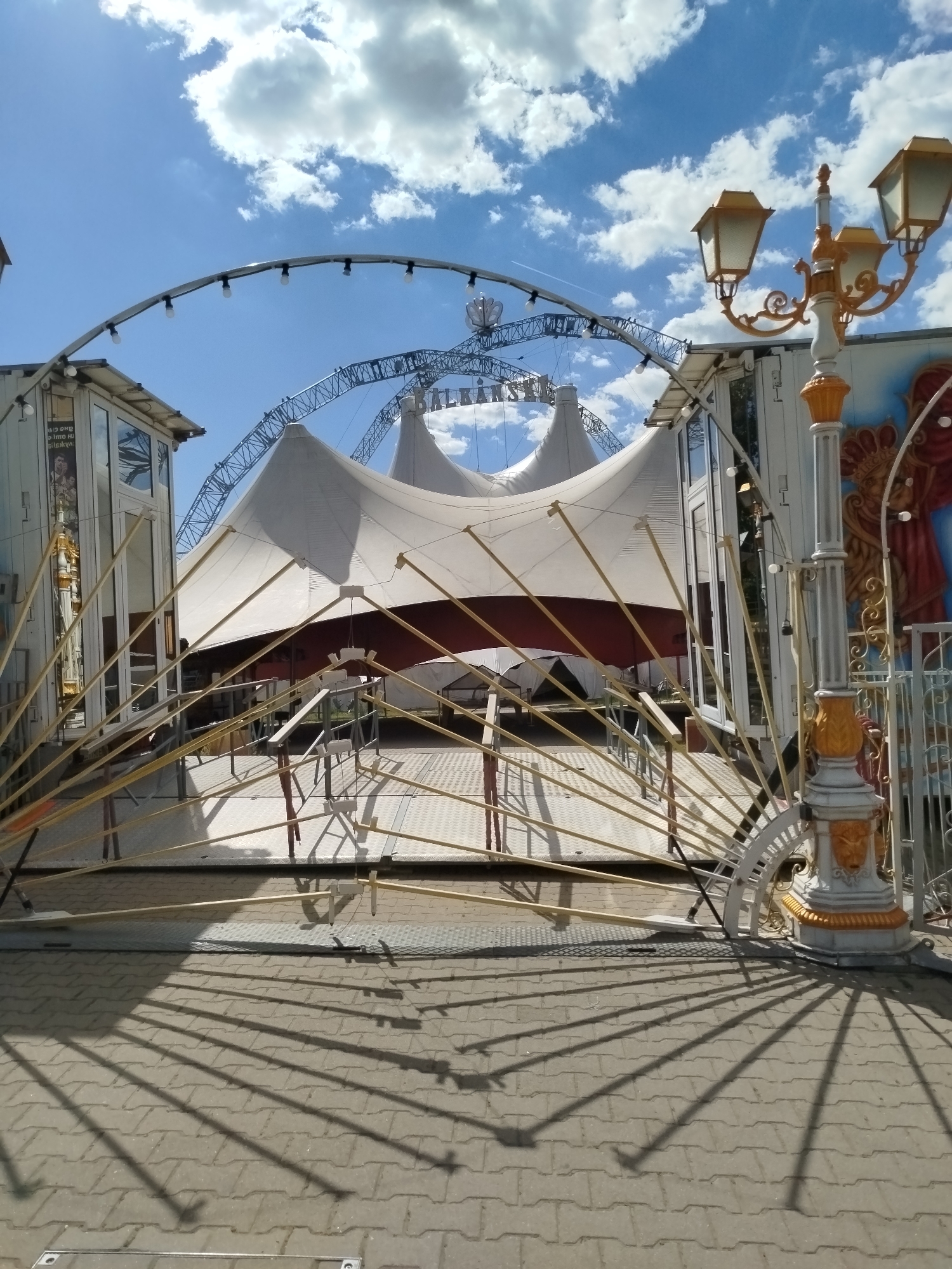 The image shows the entrance of the "Balkanski" Circus tent in Sofia, Bulgaria. The tent is white with a red horizontal stripe, and the entrance area is framed by two white structures with mirrored windows and decorated with yellow metal rods radiating outwards. A decorative street lamp stands to the right of the entrance, casting long shadows on the paved ground. The sky is blue with scattered white clouds. A metal arch with lights and the circus name is above the tent. The area appears to be devoid of people.