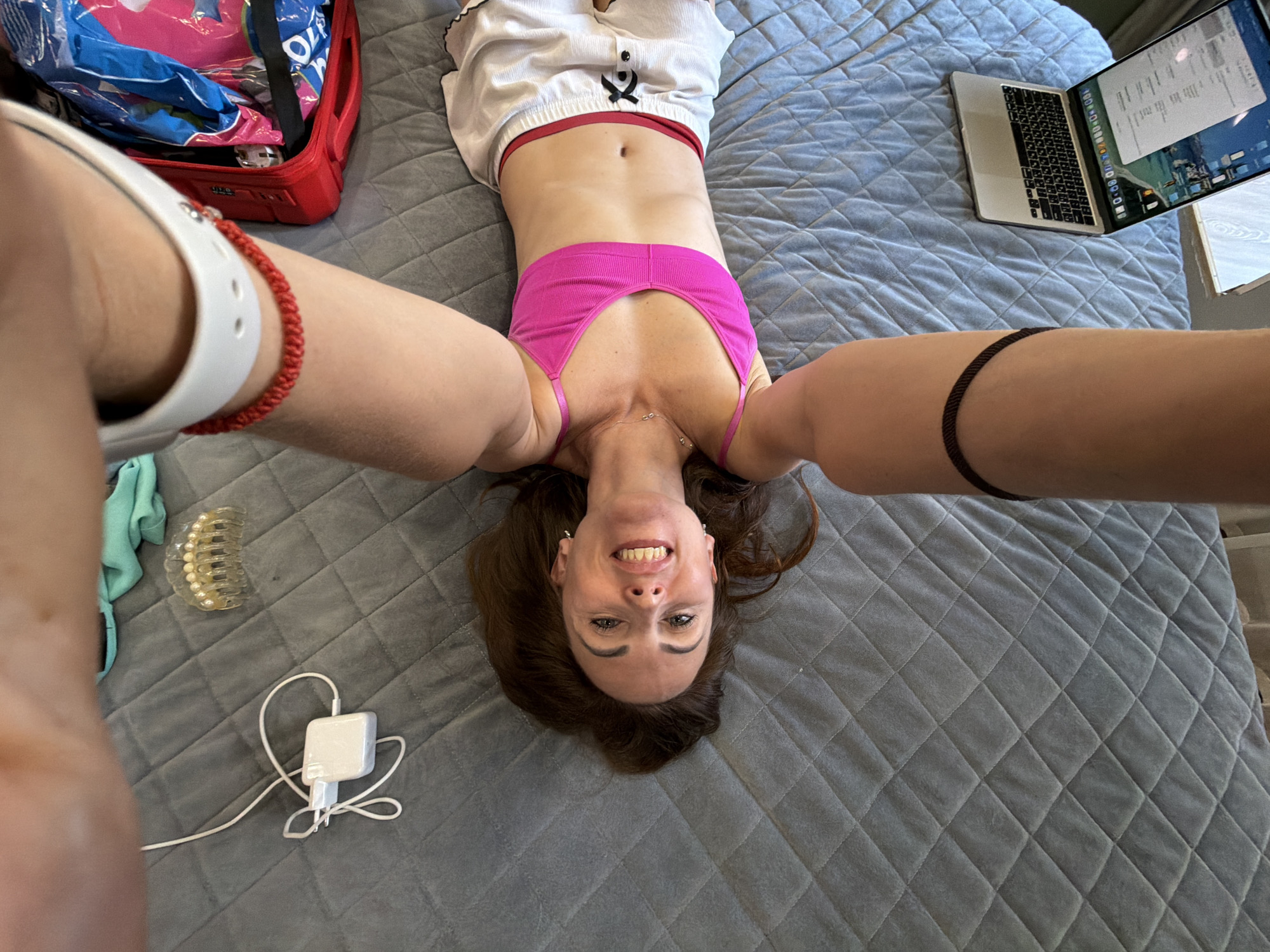 This is a high-angle, upside-down selfie of a woman lying on a grey, quilted surface that appears to be a bed. The woman has brown hair and is smiling broadly at the camera. She is wearing a pink tank top and white shorts with a red trim. Her legs are extended upwards, and her arms are also raised, holding the camera.
In the background, on the left side of the frame, is a red open suitcase filled with blue and pink items. To the right of the woman, a silver laptop is open and placed on the quilted surface. There are also a few other objects visible on the bed: a white phone charger with its cable, a mint green cloth, and a clear hair clip with pearl-like embellishments.
A thin dark bracelet is visible on the woman's right wrist and a red string bracelet is on her left wrist. A subtle necklace is visible around her neck. The lighting suggests it is daytime. The overall scene conveys a relaxed and playful mood. There is no visible text in the image. The location context provided is Birobidzhan, Russia.