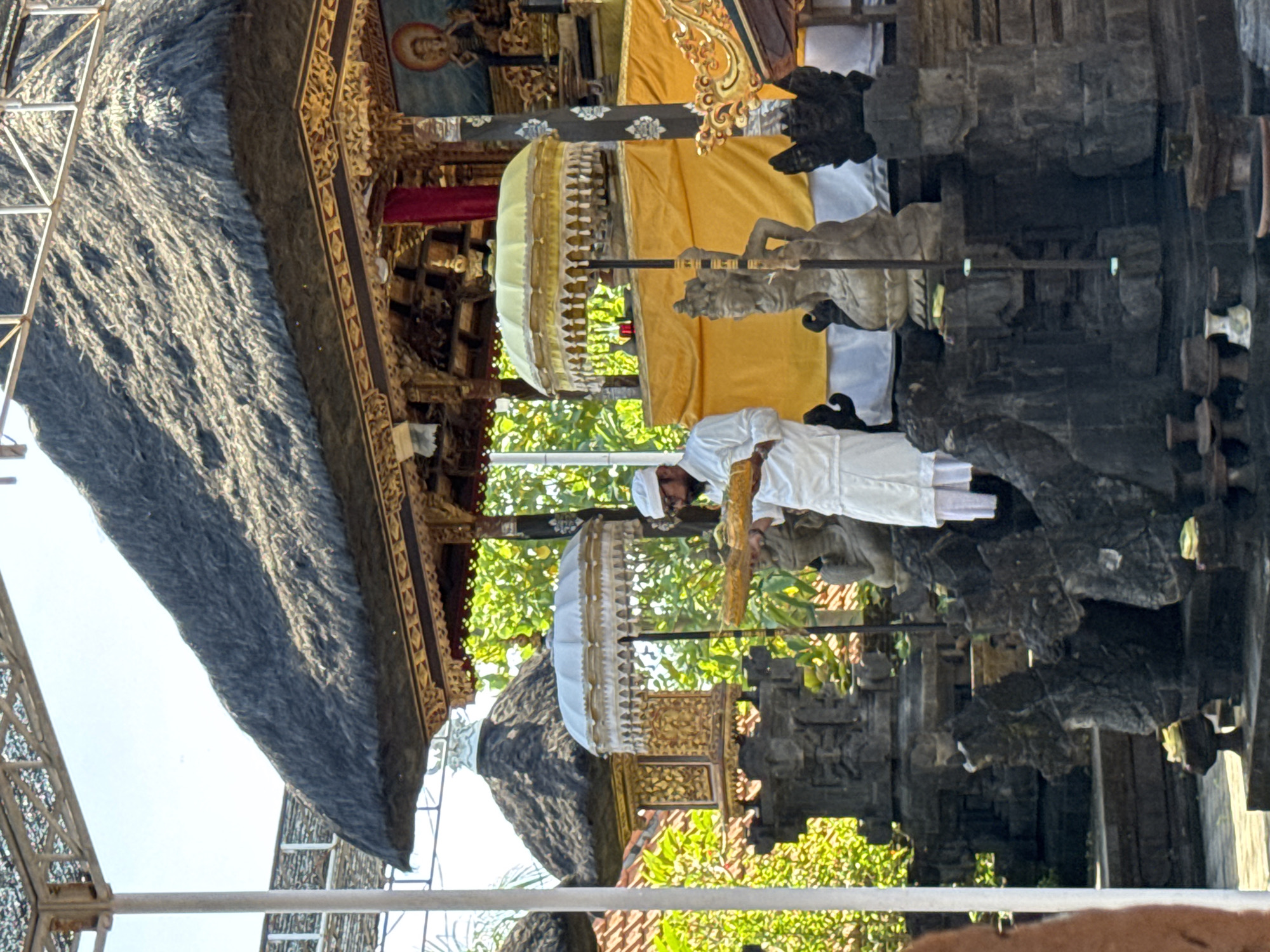 In North Kuta, Indonesia, a man in a white robe and head covering is performing a ritual at a temple. The temple features ornate carvings, a thatched roof, statues, and yellow and white ceremonial umbrellas. Foliage is visible in the background.