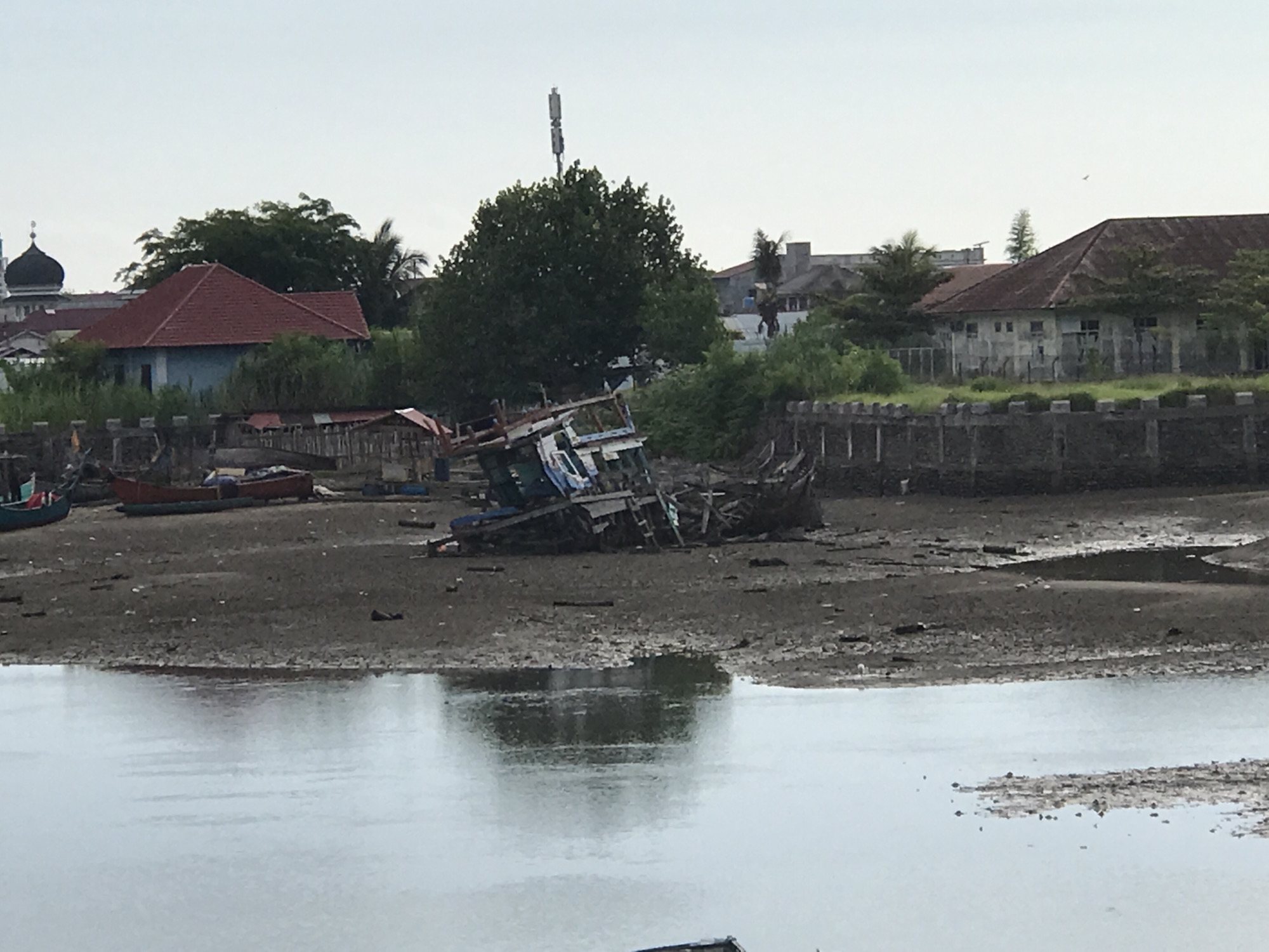 The image shows a waterfront scene in Lhokseumawe, Indonesia, featuring several boats, some of which appear to be in disrepair and grounded on a muddy shore. In the background, buildings with red tiled roofs and a mosque with a dome are visible, suggesting a settled area. There is also a prominent telecommunications tower. The water in the foreground is calm, reflecting the overcast sky. The overall impression is one of a working port or coastal community that may be experiencing low tide, leaving some of the vessels exposed.