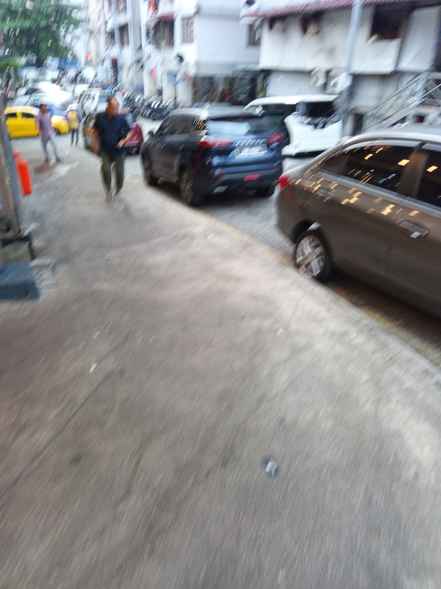 The image depicts a street scene in Kuala Lumpur, Malaysia, characterized by a blurred, dynamic motion effect. In the foreground, a person in a dark shirt and green pants is walking away from the camera. To their left, a dark blue car is parked, and further to the left, a yellow car is visible. In the middle ground, a gray car is seen from the side, with its windows reflecting the ambient light. Buildings line the street, suggesting an urban environment. The overall impression is one of a busy street, with vehicles and pedestrians in motion.