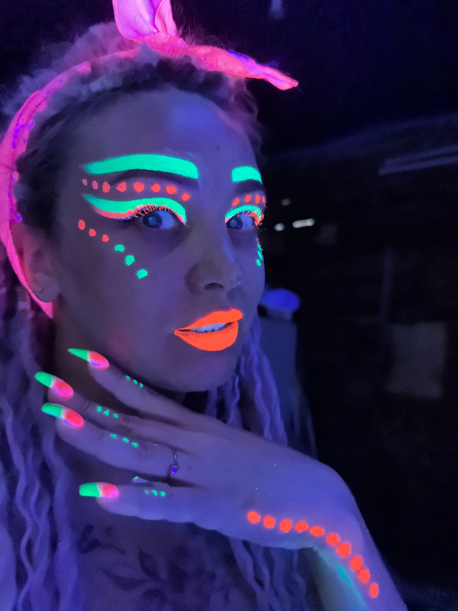 A person is under ultraviolet (UV) lighting, with glow-in-the-dark makeup applied to their face and hands. The person is wearing a pink head wrap. The individual's makeup includes green and red markings around the eyes, and bright orange lipstick. The nails are painted with a green and red design. The arm is decorated with a row of red dots. The setting is dark. There is no evidence of this photo being in Noviy, Russia.