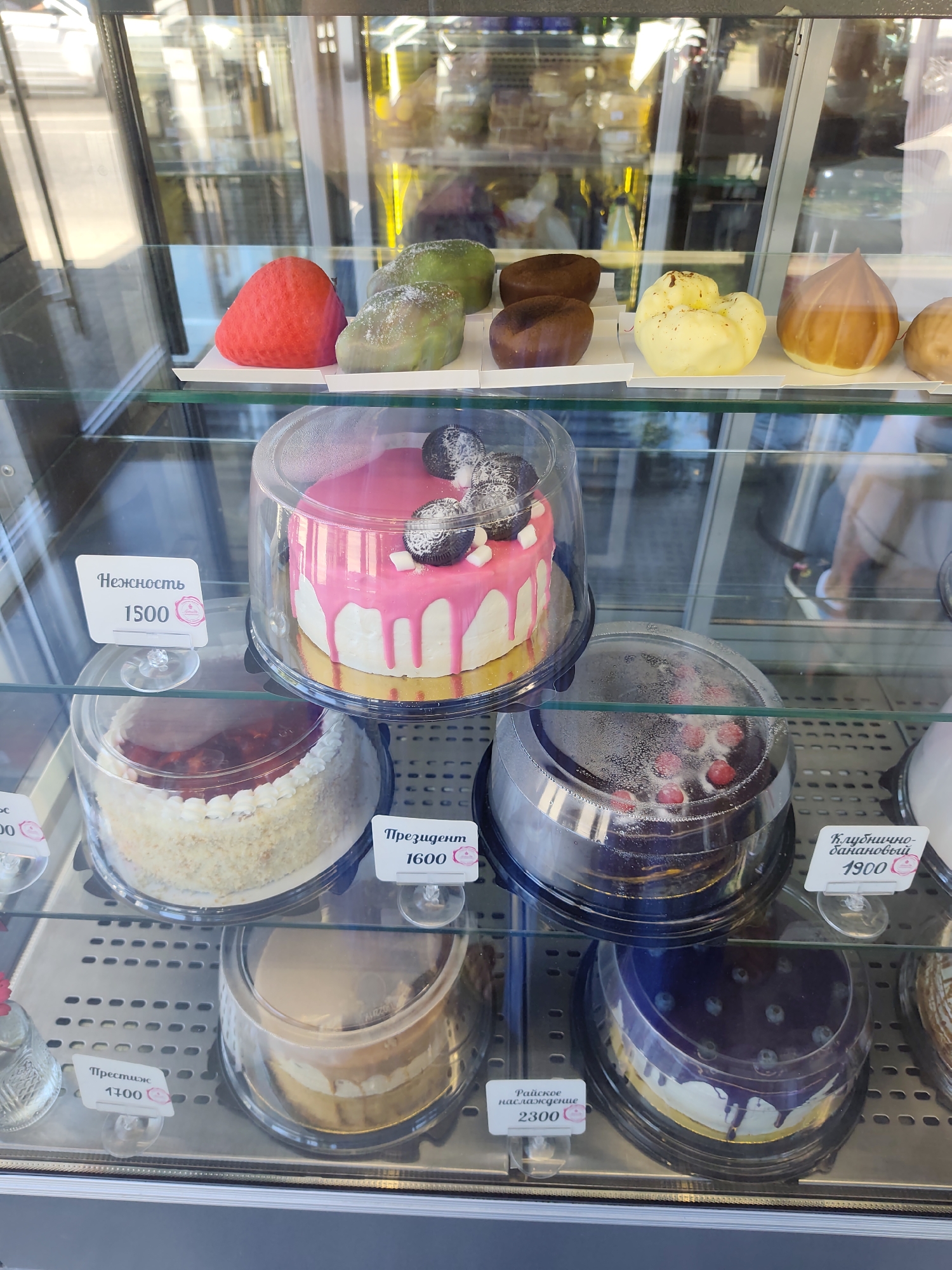 The image shows a refrigerated display case filled with various cakes and desserts in a confectionery shop. On the top shelf, several small pastries are visible, including a red strawberry-shaped one, green and brown oval-shaped ones, and a yellow one. The middle shelf features several cakes. A round cake with pink icing and white drizzles, topped with Oreo cookies and marshmallows, is prominently displayed. Next to it, another round cake with white frosting and red fruit topping is partially visible. A dark chocolate cake with red berries is also on this shelf, along with a light-colored cake with layers. The bottom shelf contains a cake with a light brown frosting and what appears to be cream layers, and a dark purple cake with blueberries and drizzles. Small price tags with Cyrillic text are placed in front of some of the items. The overall environment suggests a retail setting, likely a bakery or pastry shop. The reflection in the glass shows a blurred background, possibly a street scene with a person walking. Based on the context of the Cyrillic text and common confectionery offerings, this scene is likely located in a city within Russia, such as Sochi.