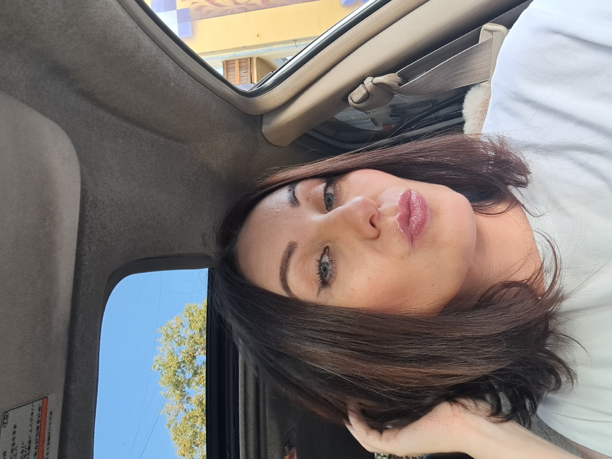 A medium shot captures a woman inside a car with her mouth pursed in a kissing gesture. She has dark brown hair, which she is touching with her left hand. Her eyes are green, and she is wearing makeup, including eyeliner and lipstick. The car's sunroof is open, revealing a blue sky and a tree. The interior of the car is visible, including the ceiling and a seatbelt. Outside the car, a building with balconies and windows can be seen. The scene appears to be a bright, sunny day.