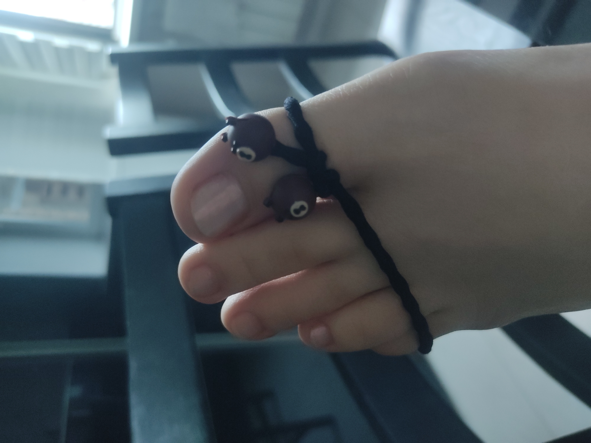 A close-up shot captures a foot wearing a black cord toe ring. The ring features two small, brown, bear-shaped beads. The foot appears to be resting on a surface with a black and gray patterned background.