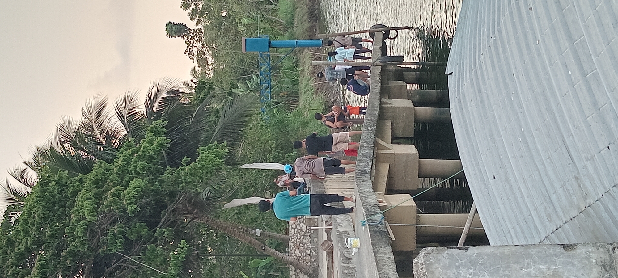 This image captures a scene at a pier in Kalipucang, Indonesia, likely during the late afternoon or early evening given the soft, diffused light. Several people are gathered on the concrete pier, which is supported by thick, cylindrical pillars extending into the calm, dark water. A large, leafy tree, possibly a palm tree, dominates the left side of the frame, its branches extending over the pier. Dense green foliage forms the background.

On the pier, a group of individuals are engaged in various activities. A person in a teal shirt stands facing away from the camera, while others are seen standing, sitting, and interacting. One person, wearing a blue cap, appears to be talking to another. A man in shorts and a t-shirt is sitting. Further down the pier, a couple of individuals are observing something in the water. A child in an orange dress is standing near the edge. The pier itself is aged, with visible cracks and a slightly uneven surface. A blue cylindrical structure with a metal frame is visible in the background, possibly part of a navigation aid or infrastructure. In the foreground, a corrugated metal roof curves across the bottom of the frame, suggesting it belongs to a nearby structure. The water reflects the muted tones of the sky and surrounding vegetation.