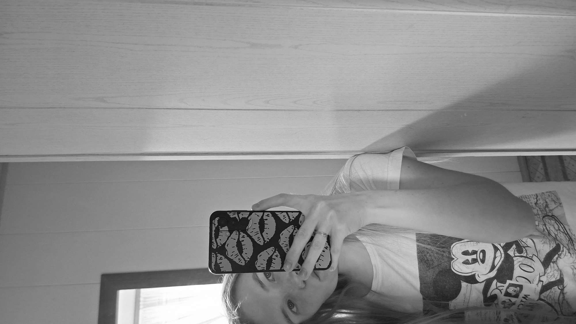 A young woman is taking a selfie in a mirror. She has blonde hair and is wearing a t-shirt with Mickey Mouse printed on it. Her phone has a black case with a pattern of lips. She is wearing a ring on her left hand. The background appears to be a room with wooden paneling and a closet. The image is in black and white.