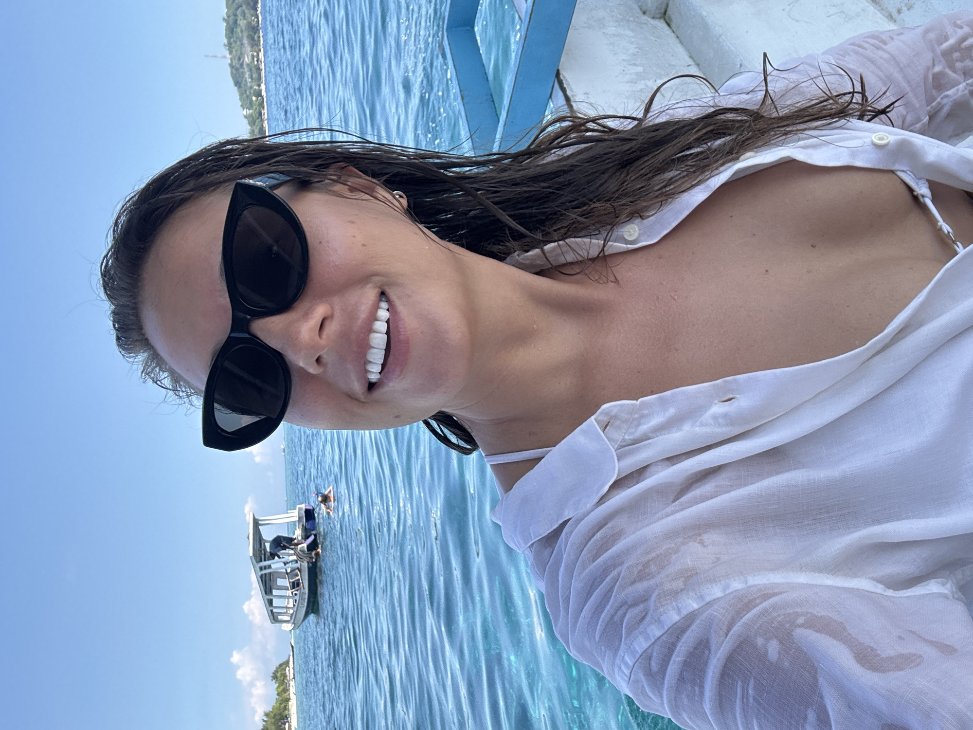 A young woman is seen in a medium shot, seemingly taking a selfie on a boat in Unknown, Indonesia. She is smiling, wearing large black sunglasses and a wet, partially unbuttoned white shirt over what appears to be a white bathing suit. Her hair is wet. The bright blue ocean is behind her, with another small boat visible in the distance to her left. The sky is blue with some wispy clouds. The angle is slightly off-kilter, suggesting a handheld shot.