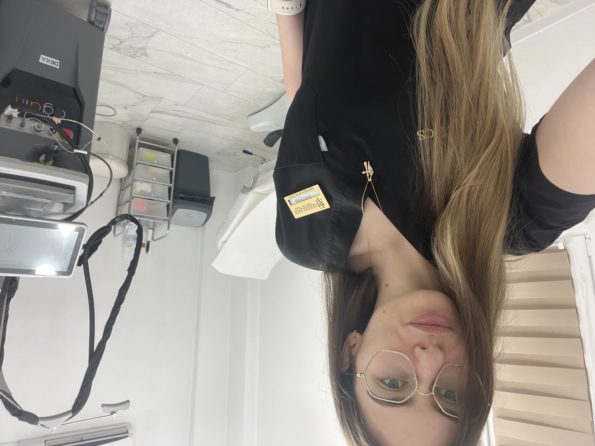 A woman with long, blonde hair, wearing gold-rimmed glasses and a black uniform, is centrally positioned in an indoor medical setting. She has fair skin and green eyes, and a gold necklace with the letters "SD" is visible. The black uniform features a name tag and a small, decorative brooch or pin.
The background features medical equipment, including a DEKA-branded machine on wheels. There is a white examination chair and a multi-tiered storage unit with clear drawers and a dark gray container next to it. The walls and floors are light-colored, giving the space a sterile appearance. The specific location in the city of Unknown, Unknown, cannot be ascertained from the image.