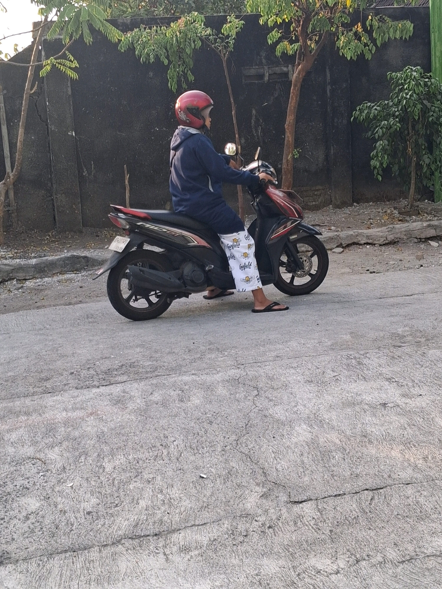 An individual wearing a red helmet and a dark blue hoodie is seated on a black and red scooter on a paved road. The person is wearing white pants with a black Garfield cartoon print. The scooter has a license plate with the characters "D4DX." The background features a dark, textured wall and several trees. The environment appears to be an outdoor urban setting, likely in Surakarta, Indonesia, given the context of the surrounding area.