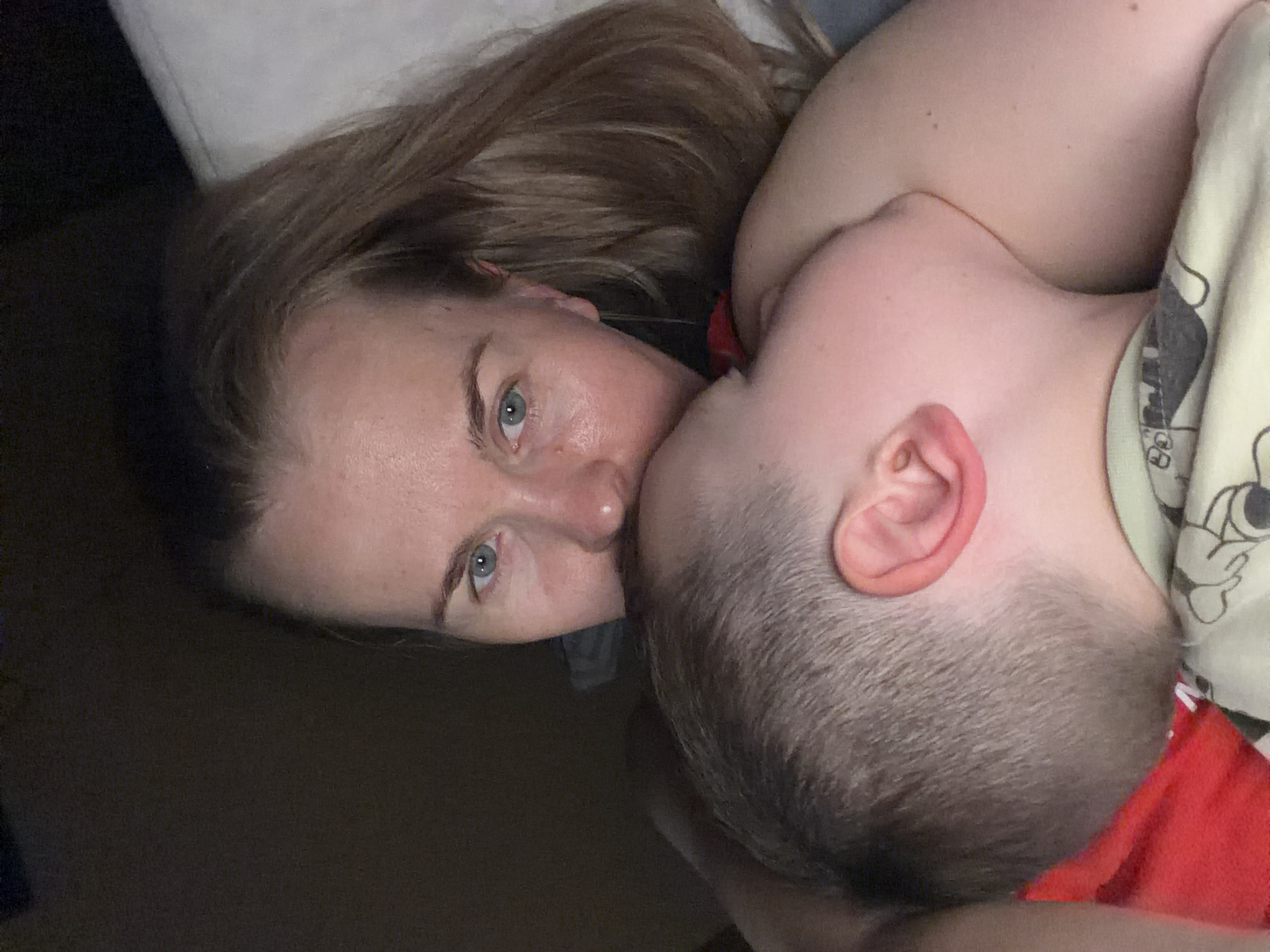 A low-angle, close-up, indoor shot shows a woman holding a young child. The woman's face is turned towards the camera, with her blue eyes looking directly at the viewer. Her blonde hair is long and falls over her shoulder. The child is nestled into the woman's chest, with their head turned away from the camera. The child's ear is visible, as is the back of their head. The background is dark and indistinct, with a hint of a white surface on the right side, possibly a pillow or mattress. The woman is wearing a grey shirt, and the child is wearing a green shirt with a pattern. The image appears to have been taken at night or in a dimly lit room. There is no identifiable information in the image to suggest a specific location within the city of Khabarovsk, Russia.