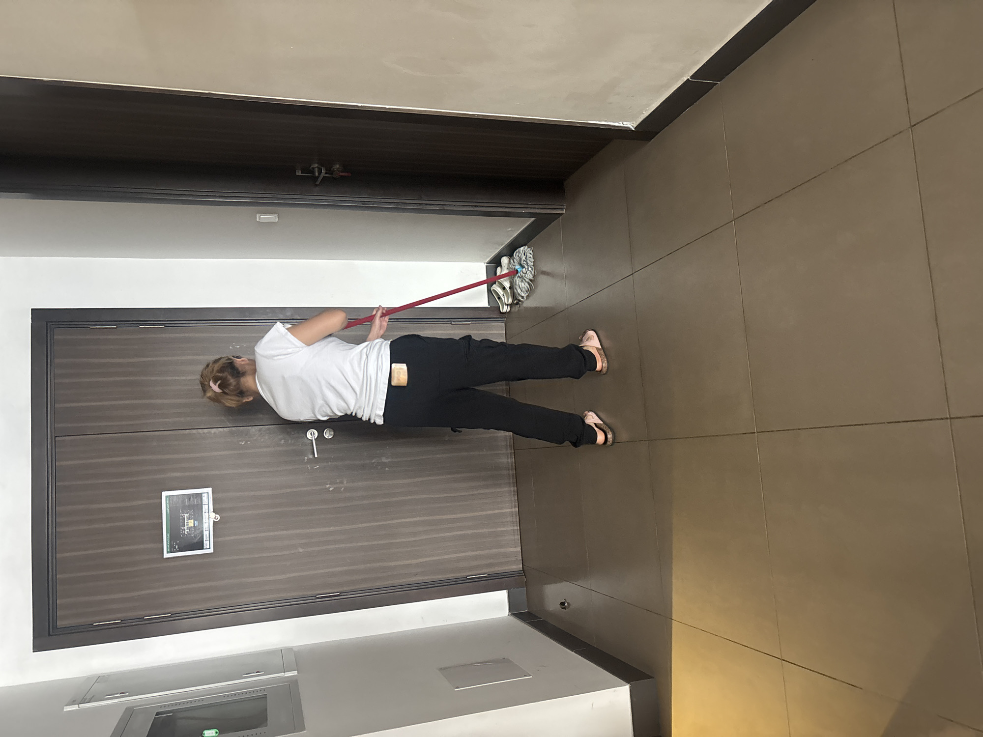 A person is mopping a tiled floor in a hallway. The person, viewed from the back, wears a white t-shirt and black pants. They are holding a red-handled mop with a white mop head, which is wet. Two pairs of sandals are near the mop. The hallway has large, square, brown tiles on the floor and cream-colored walls. There are two dark wood doors in the background, one with a sign attached. To the left, a grey electrical panel is mounted on the wall. The scene appears to be indoors, possibly in an apartment building corridor.