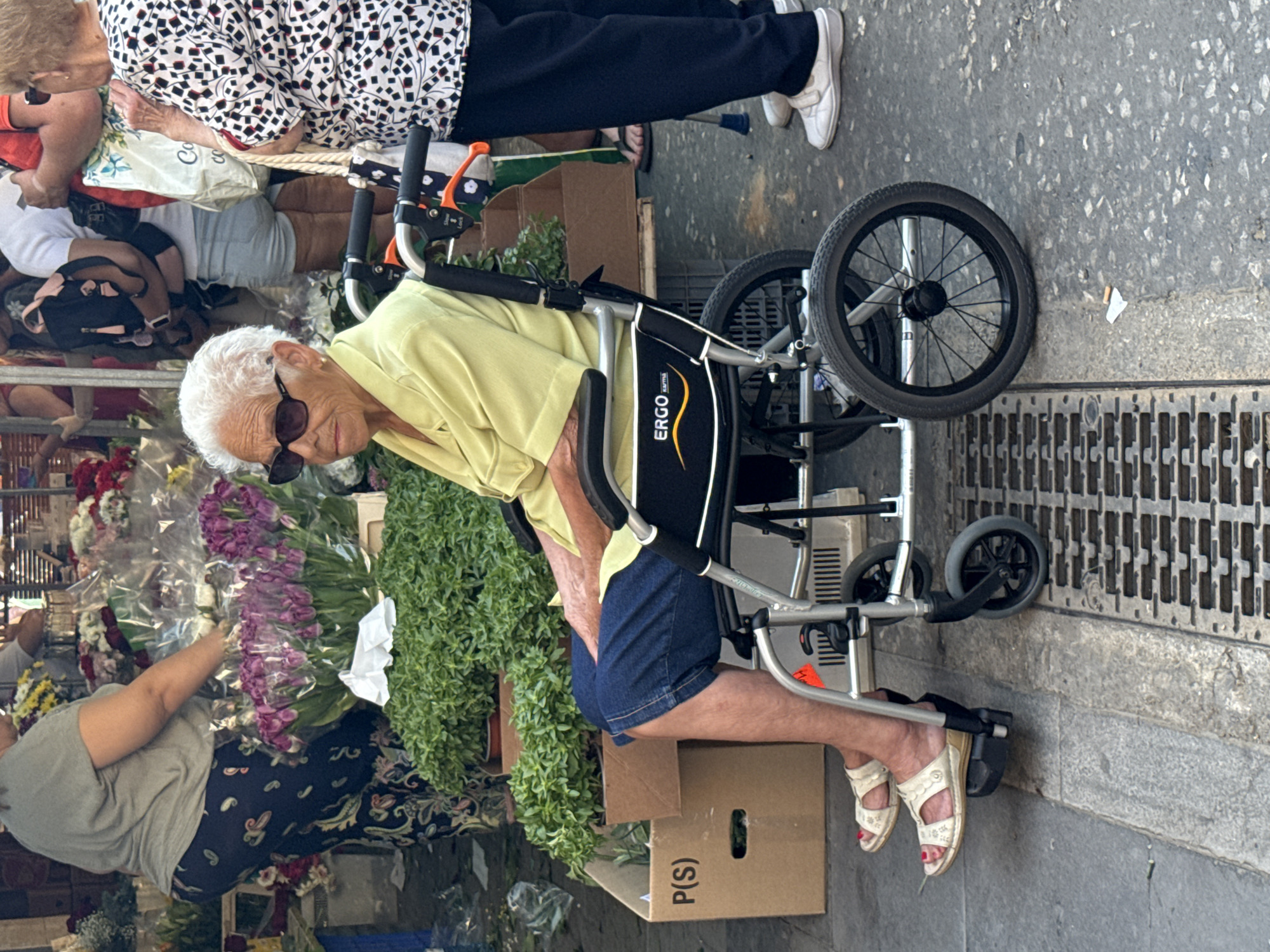 In an outdoor urban setting in the city of Unknown, Unknown, an elderly woman is positioned in a silver Ergo Karma walker. She is wearing a light yellow shirt, denim shorts, and white sandals with visible red toenail polish. The woman is also wearing sunglasses and has white hair. She is amidst an apparent outdoor market, specifically near a flower stall with various flowers including tulips and greenery. Other individuals, presumably shoppers, are visible in the background, wearing diverse attire such as black and white patterned tops, black pants, white tops, and dark patterned pants. Boxes labeled "P(S)" are situated near the walker, and the ground is paved. A metal grate is seen adjacent to the walker's wheel.