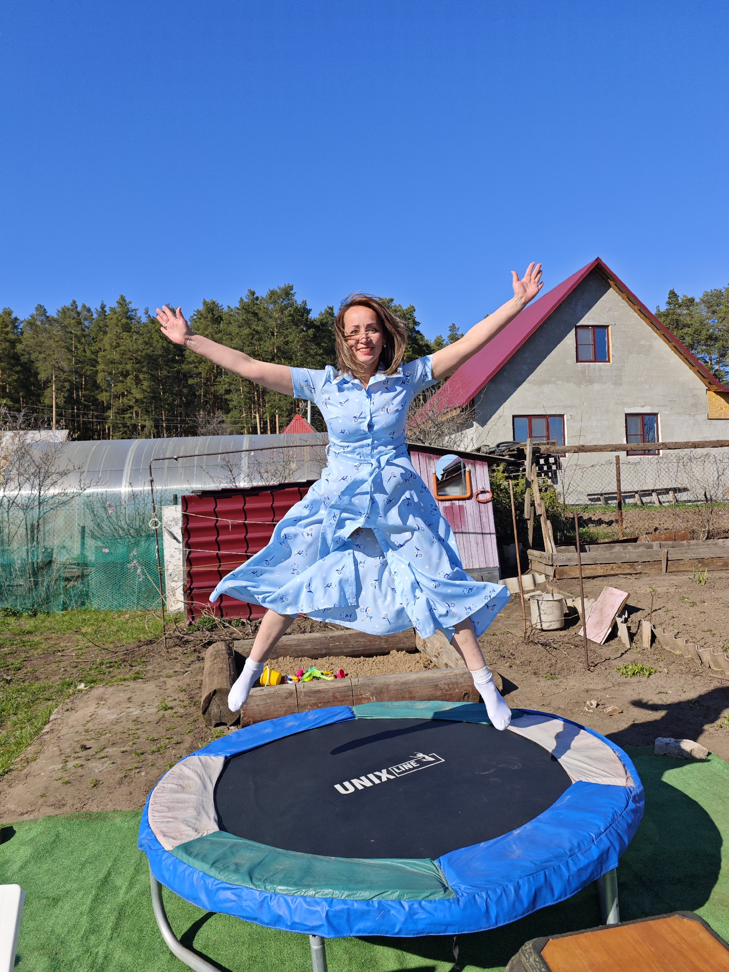 A woman in a blue dress with a bird print is jumping on a round trampoline with a blue and green padded rim in a backyard. She is wearing white socks and has her arms outstretched. The trampoline has the words "UNIX LINE" on its black surface.
Behind the trampoline, there is a wooden structure, a greenhouse, and a gray house with a red roof. A forest line is visible in the background under a clear blue sky. The setting is in a garden with various other items scattered around, like gardening tools and wooden structures.