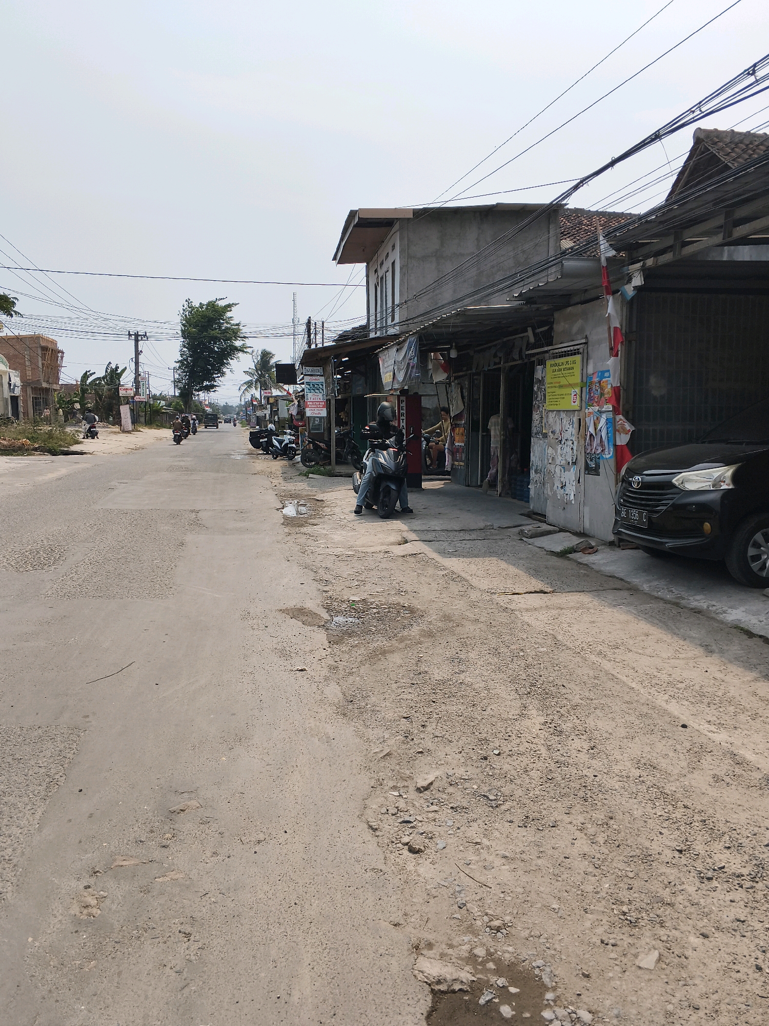 This is a medium-angle, full shot of a street scene in Bandar Lampung, Indonesia, on a clear, sunny day. The street is paved but appears to be in disrepair with visible patches and potholes. On the right side of the street, a row of single-story buildings lines the road, appearing to be small shops or businesses. These buildings have corrugated metal roofs and open fronts, revealing merchandise and activities within. A black Toyota Avanza car with license plate BE 1356 C is parked in front of one of these establishments. Several motorcycles are parked and in use on the street. On the left side of the street, there are more buildings under construction, some with visible brickwork. Electrical wires are strung overhead, supported by utility poles. A few trees are scattered throughout the scene, with a taller, leafy tree prominent in the mid-ground. The overall impression is of a moderately busy urban street with a mix of commercial activity and ongoing construction.