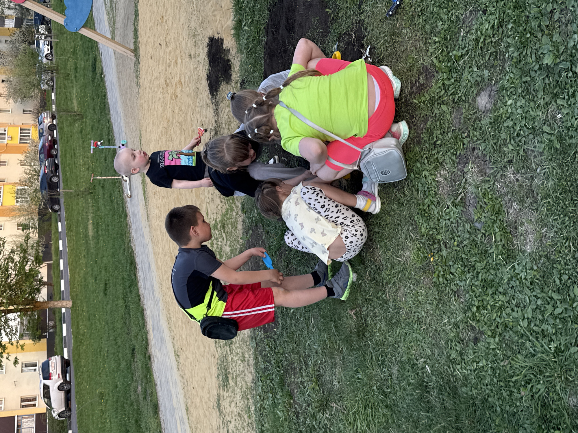 The image depicts a group of children playing in an outdoor area in Severnyi, Russia. The children, of varying ages, are gathered near a patch of dirt surrounded by grass. Some are kneeling or crouching, appearing to be digging or playing with the dirt. A young boy is seated on the grass, holding a blue object. Another child stands nearby, holding a red object. A sandbox and playground equipment are visible in the background. Buildings and cars are also seen in the background, indicating an urban setting.