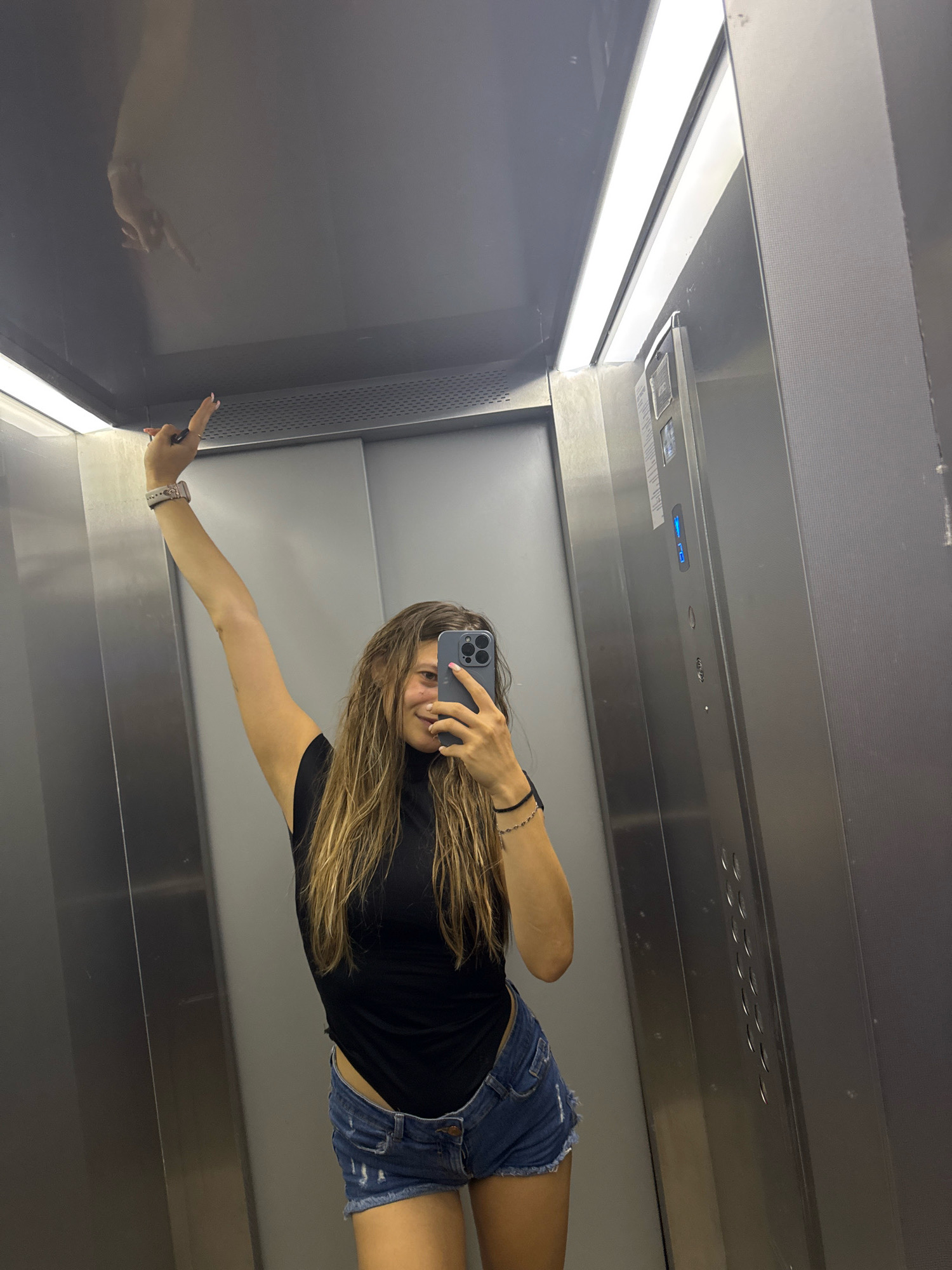 A young woman with long, light brown hair is taking a selfie in an elevator. She is wearing a black sleeveless top and denim shorts. Her left arm is raised above her head, and she is holding her phone in her right hand to take the picture. The elevator is made of brushed metal, and the doors are closed. There are fluorescent lights on the ceiling, and a panel of buttons is visible on the right wall. The woman is smiling and appears to be in a playful mood. The reflective surfaces of the elevator create a distorted image of her hand reaching towards the ceiling. The image quality is average, with some noise visible. It is not possible to determine the exact location in the city of Sofia, Bulgaria from the image alone.