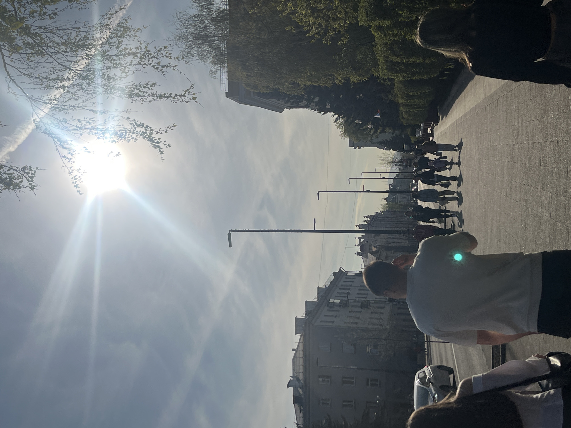 The image shows a bright, sunny day in a city with European-style architecture. A group of people is walking in the same direction along a wide pedestrian path. The camera is located at ground level looking up. The buildings appear to be apartment blocks. There are trees and lampposts lining the street. The location is Krasnoyarsk, Russia. A man in a white t-shirt and dark shorts stands in the foreground facing forward.
