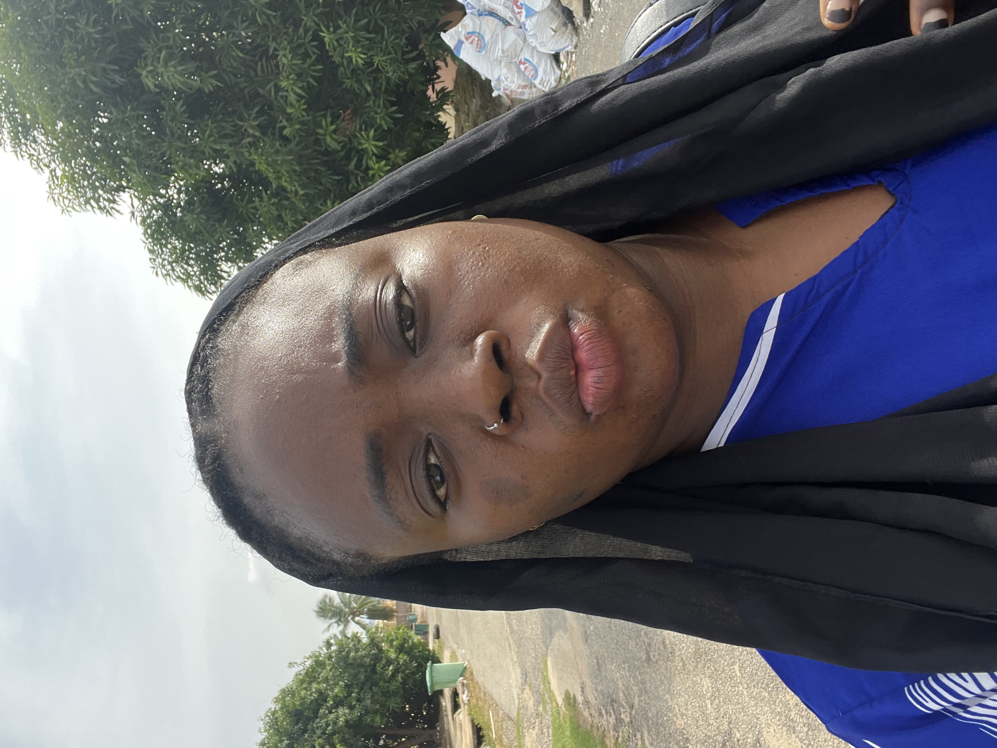 A medium, close-up shot shows a dark-skinned woman in a blue shirt with white stripes and a black head covering. She has a nose ring in her left nostril, small gold earrings, dark eyes, and pink lips. The background suggests an outdoor urban environment, possibly in the city of Abuja, Nigeria, with trees and buildings visible. In the upper left, a green tree is visible and in the upper background, white sacks are stacked. The woman has nails with black nail polish and white tips. The lighting is bright and it appears to be daytime.